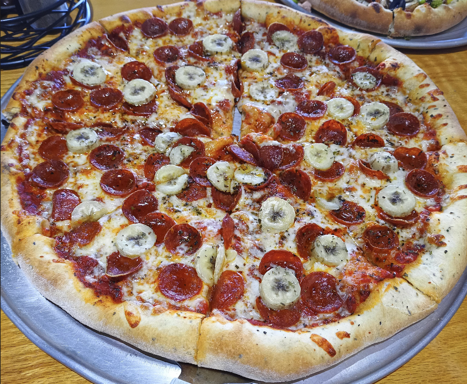 A large pizza with pepperoni and banana slices as toppings, sitting on a metal tray atop a wooden table. The pizza is sliced and has a golden, thick crust.