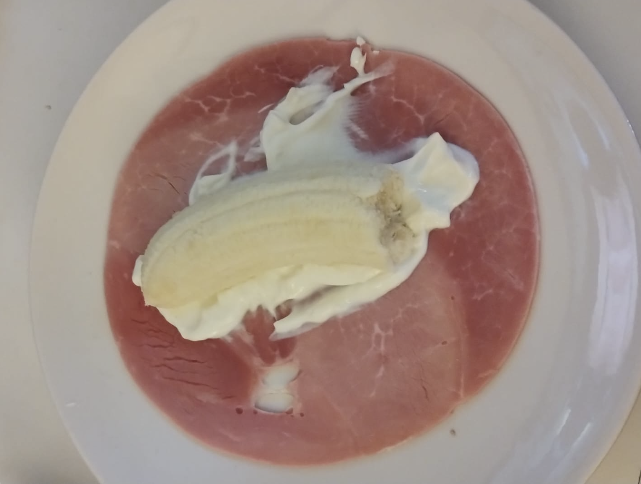 A peeled banana on a dollop of white cream sits atop a slice of pink ham, all arranged on a white plate.