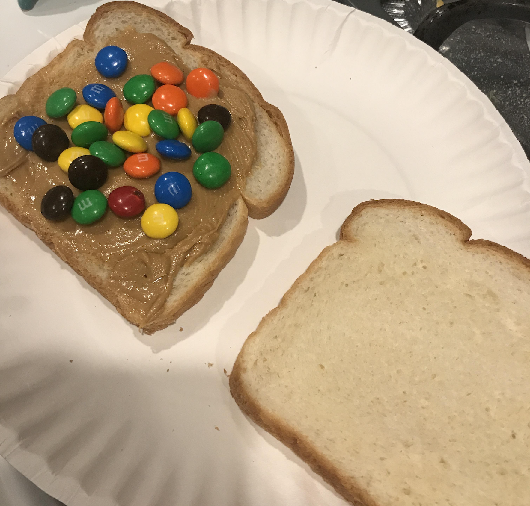 Two slices of white bread on a paper plate. One slice has peanut butter spread on it, topped with colorful M&M candies. The other slice is plain.