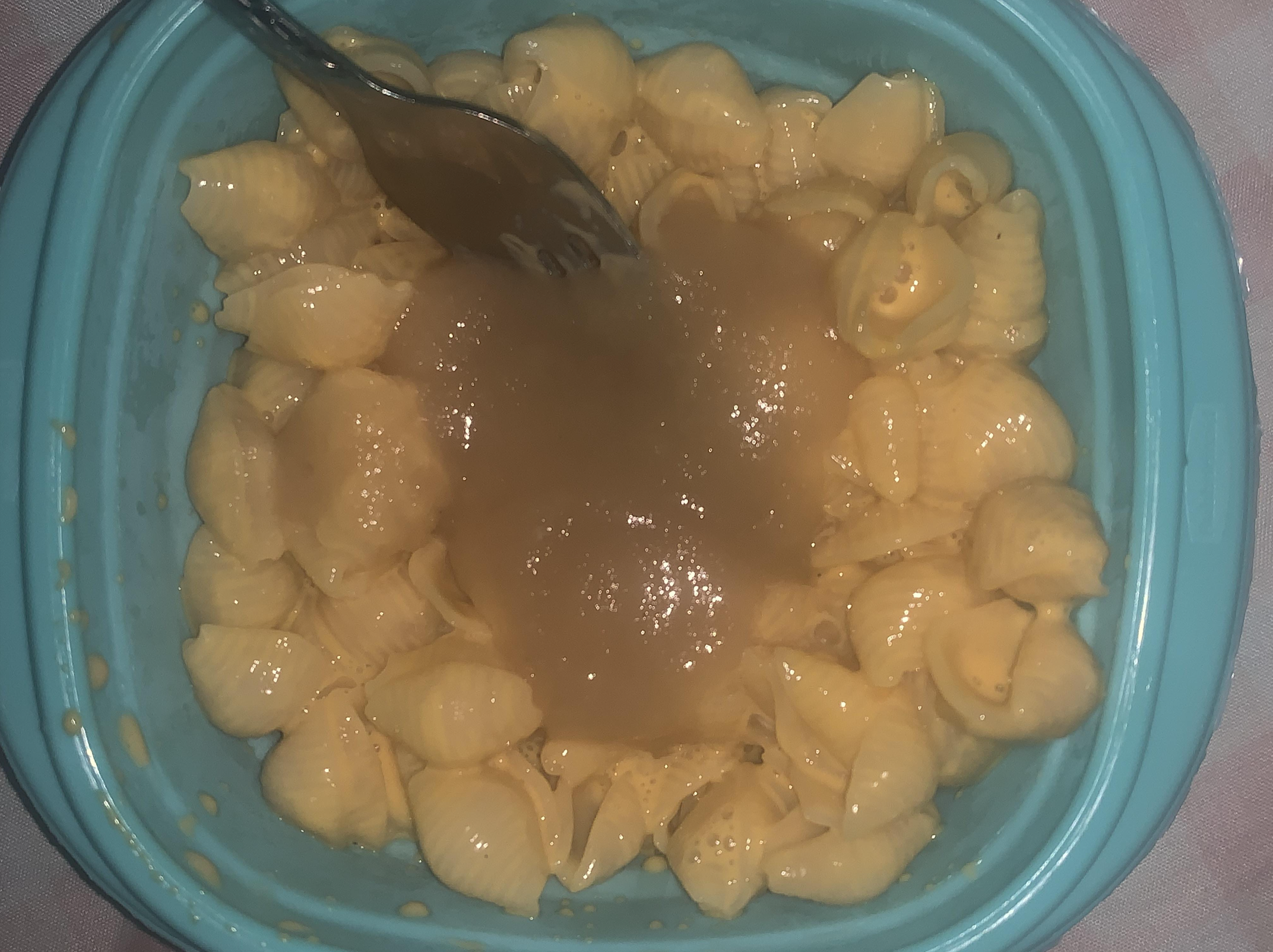 A blue bowl filled with shell macaroni and cheese topped with a heap of applesauce, with a fork resting on top.