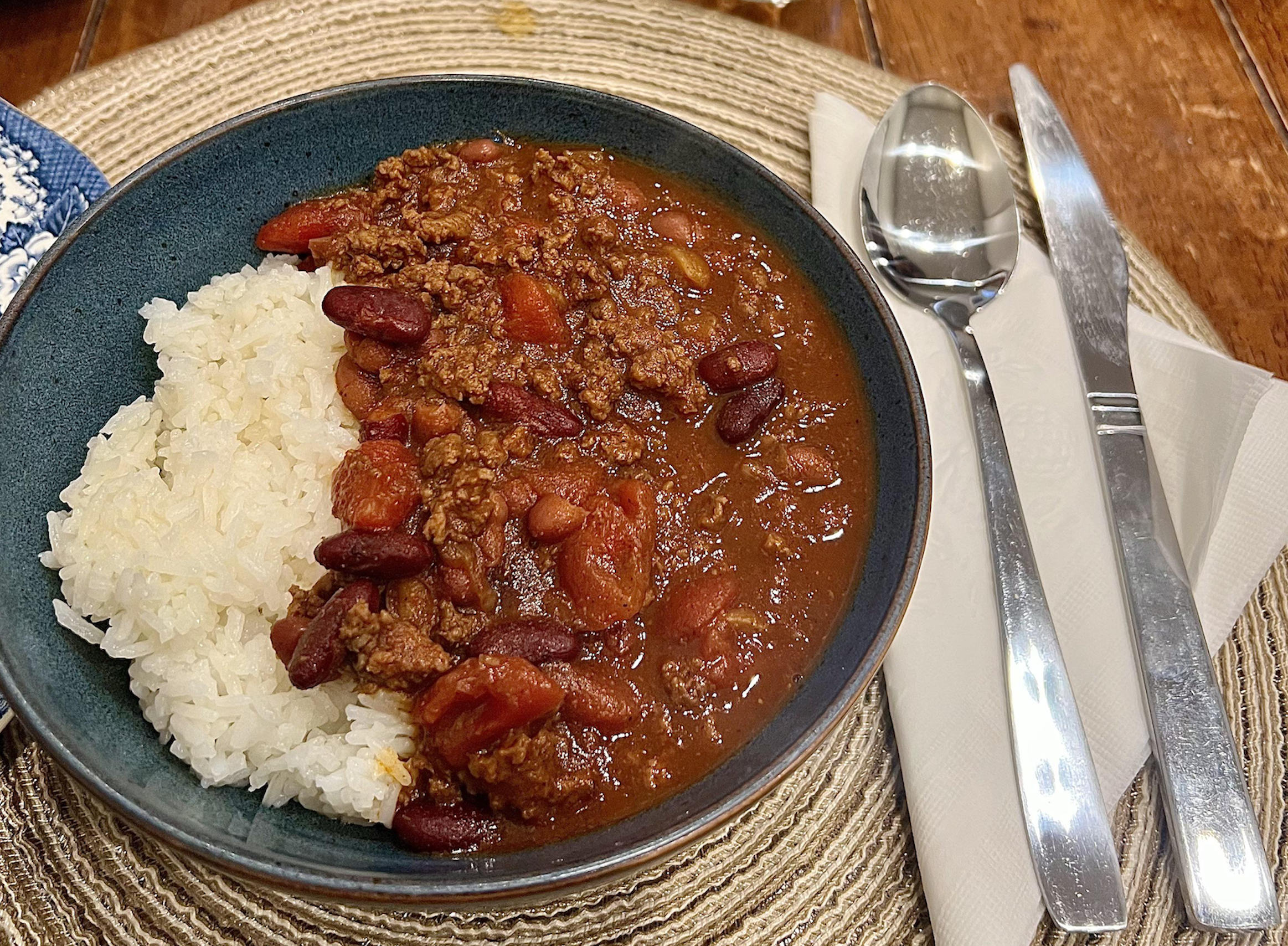 A blue bowl with white rice and chili with ground beef, kidney beans, and tomatoes, on a woven placemat. A fork, knife, and spoon are placed on a napkin beside the bowl.