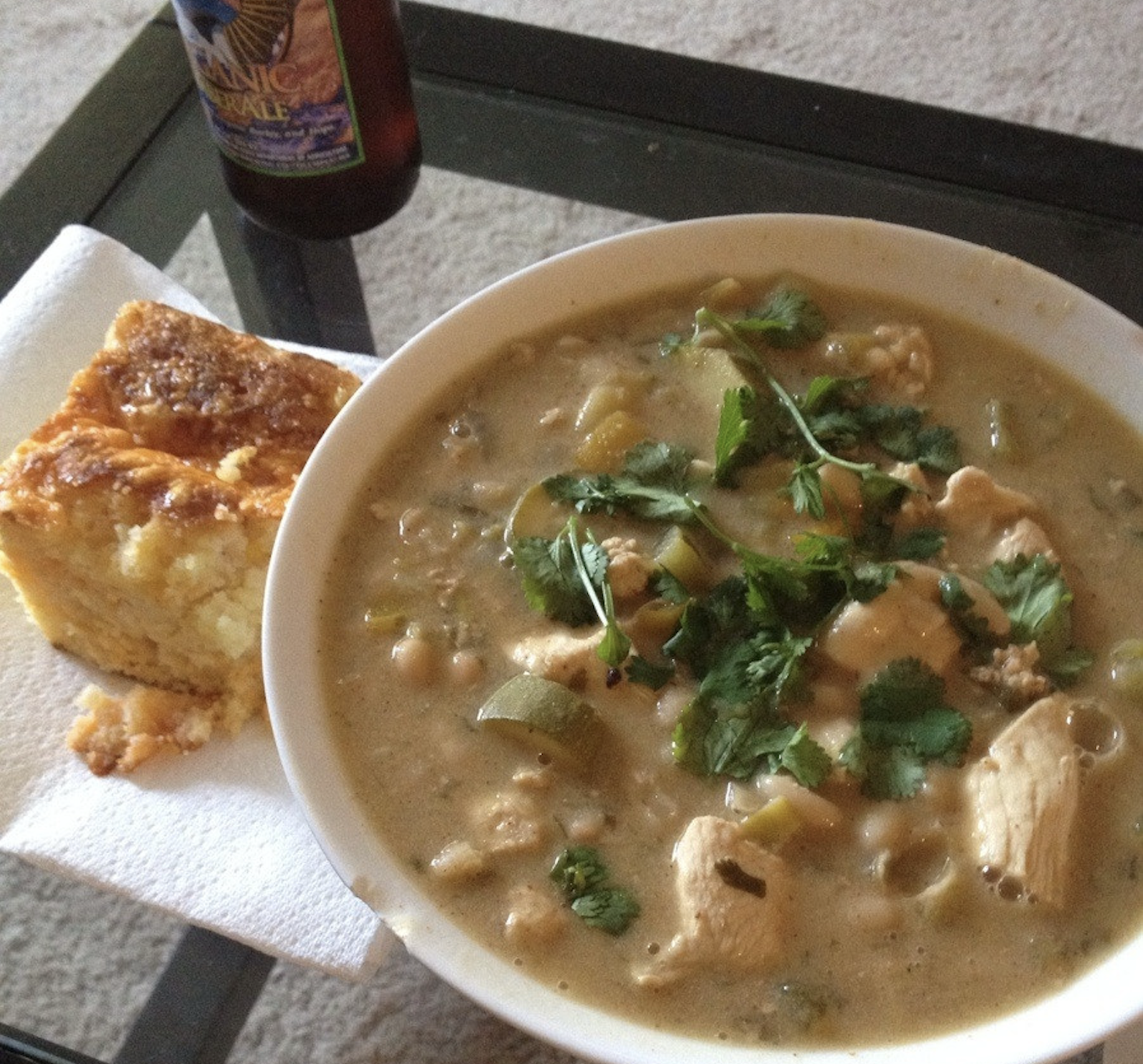 A bowl of chicken chili topped with cilantro sits on a table beside a piece of cornbread on a napkin. A bottle of beer is partially visible in the background.