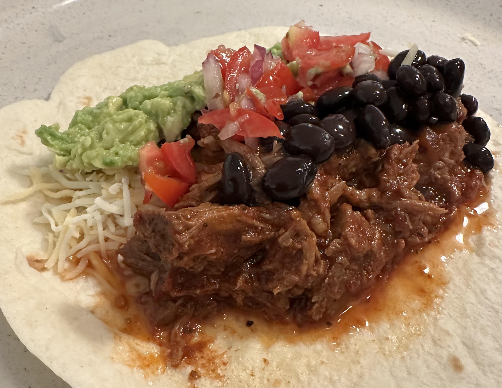 A flour tortilla topped with shredded beef in sauce, black beans, shredded cheese, guacamole, and fresh salsa with diced tomatoes, onions, and cilantro.