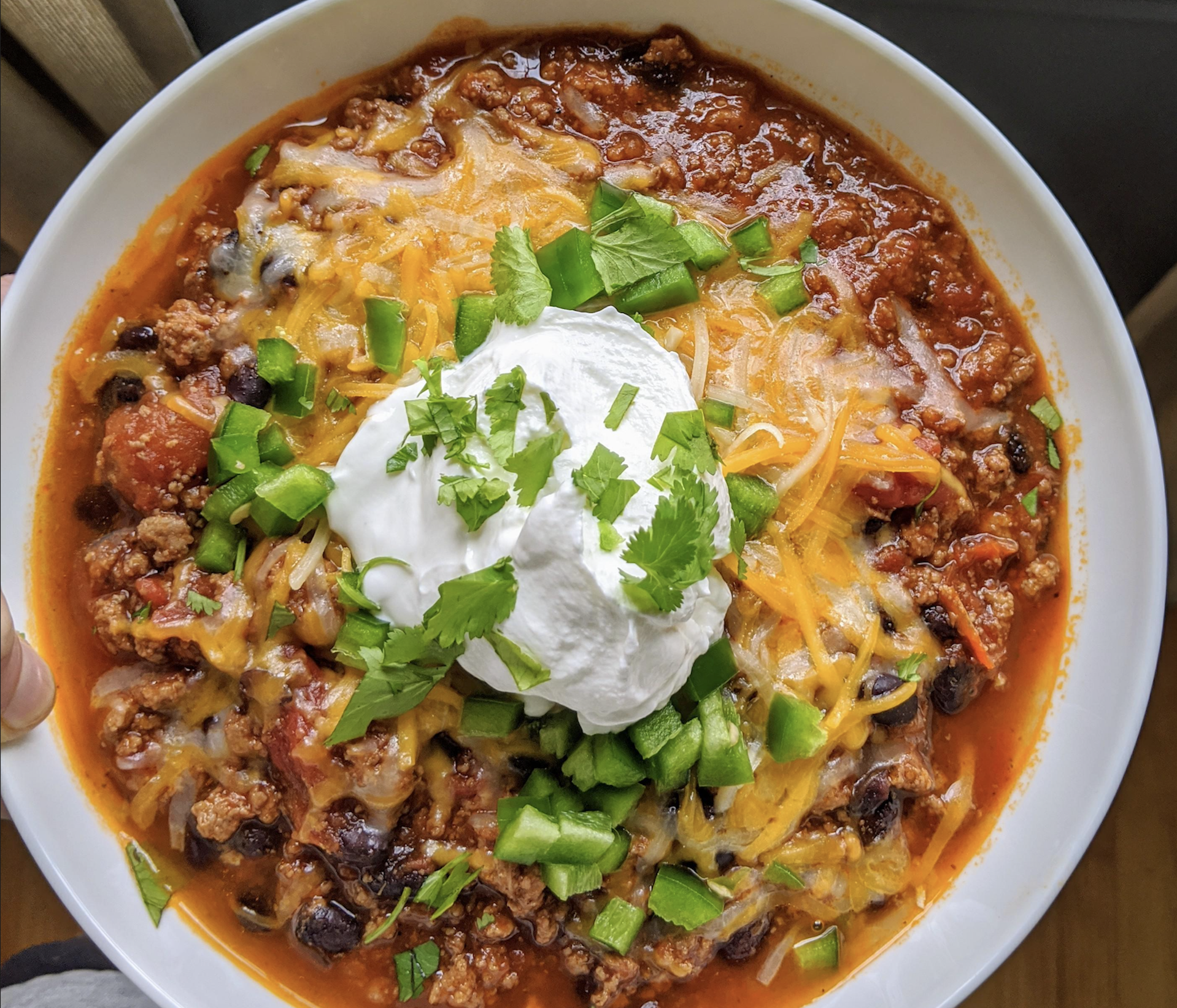 A bowl of chili topped with shredded cheese, sour cream, chopped green peppers, and cilantro. The chili contains ground meat, black beans, and tomatoes.