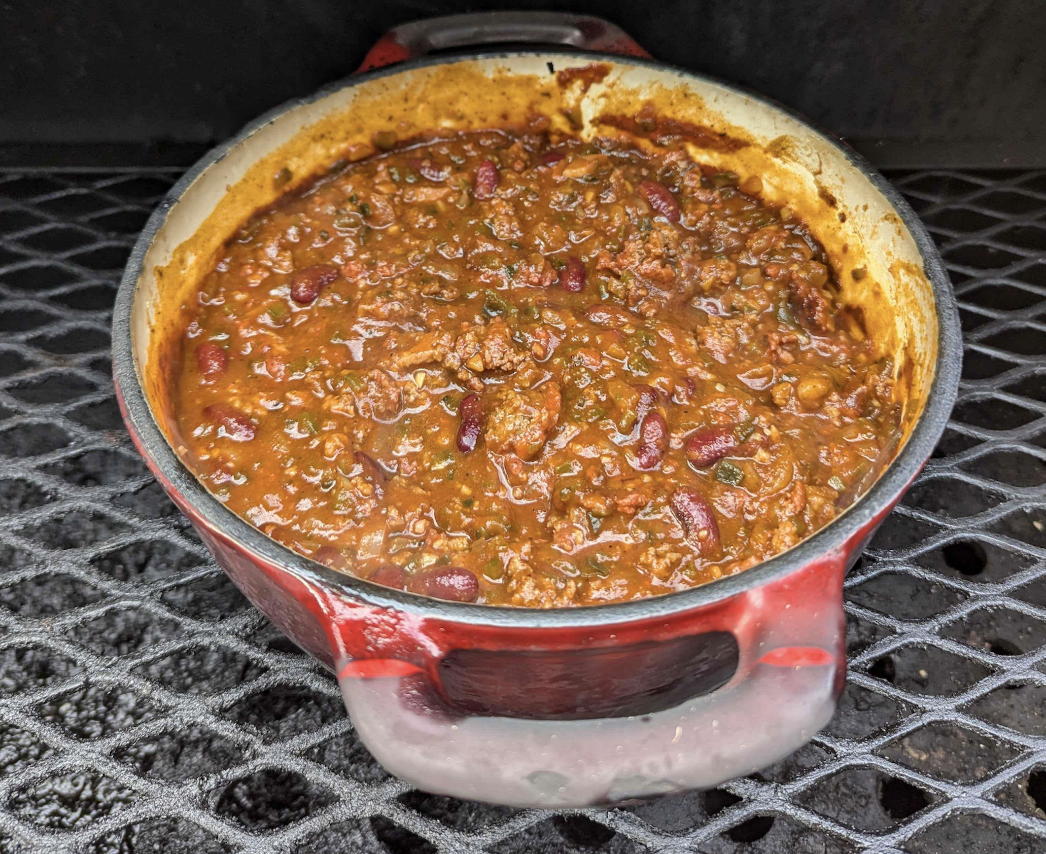 A red pot filled with chunky chili, featuring ground meat, beans, and tomatoes, sits on a metal grill with a dark background.