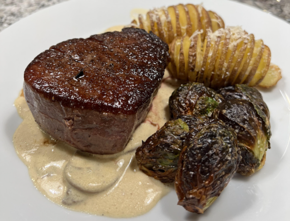 A seared filet mignon sits on creamy sauce, served with crispy Brussels sprouts and sliced roasted potatoes topped with grated cheese on a white plate.
