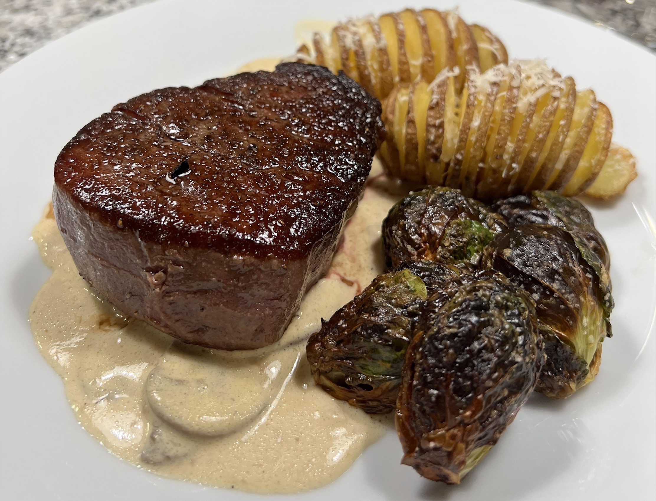 A seared filet mignon sits on creamy sauce, served with crispy Brussels sprouts and sliced roasted potatoes topped with grated cheese on a white plate.