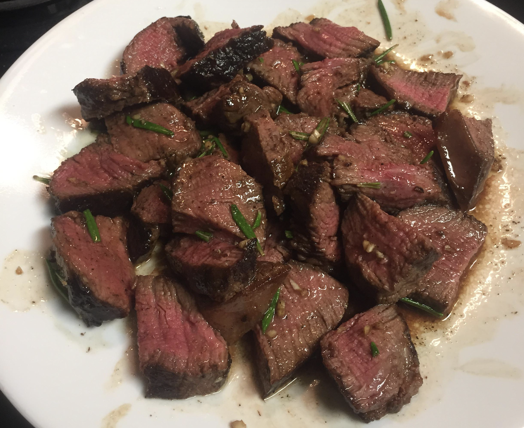 Chunks of seared steak, medium-rare with a pink center, garnished with fresh herbs, served on a white plate with some juices visible.