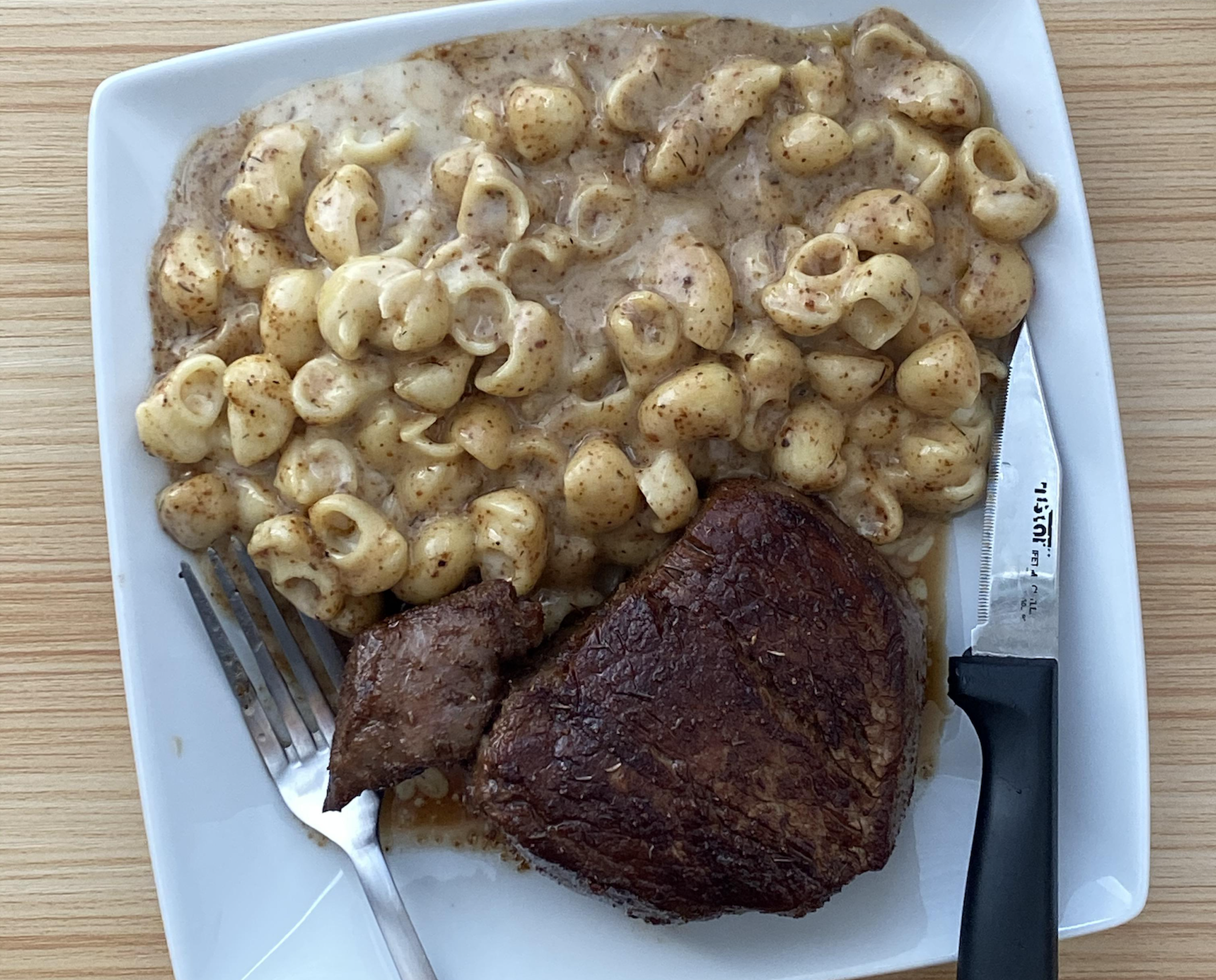 A square white plate with macaroni and cheese in sauce next to a grilled steak. A fork and steak knife rest on the plate. The table is light wood.