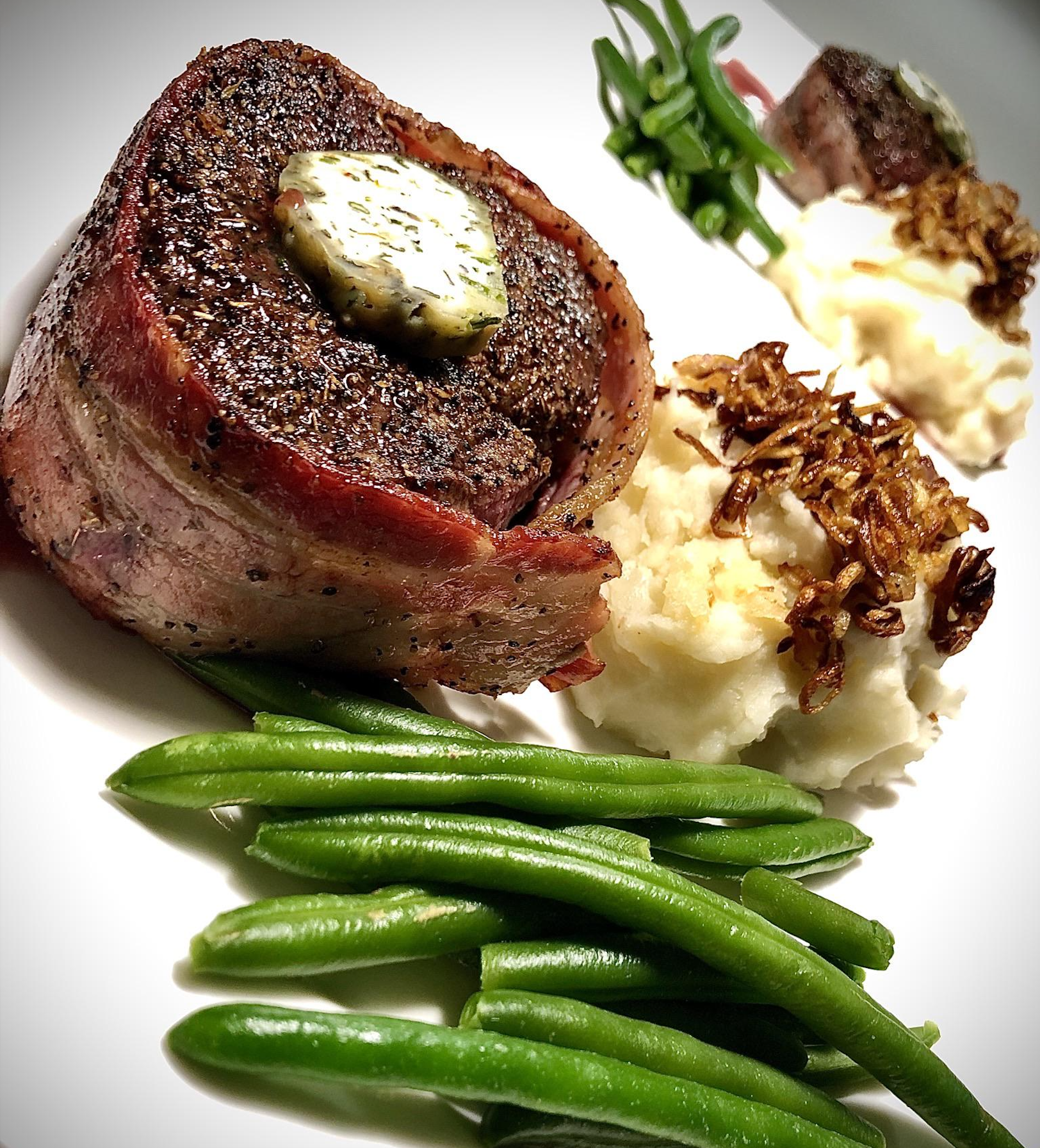 Filet mignon wrapped in bacon with herb butter, served with mashed potatoes topped with crispy onions, and green beans on a white plate.