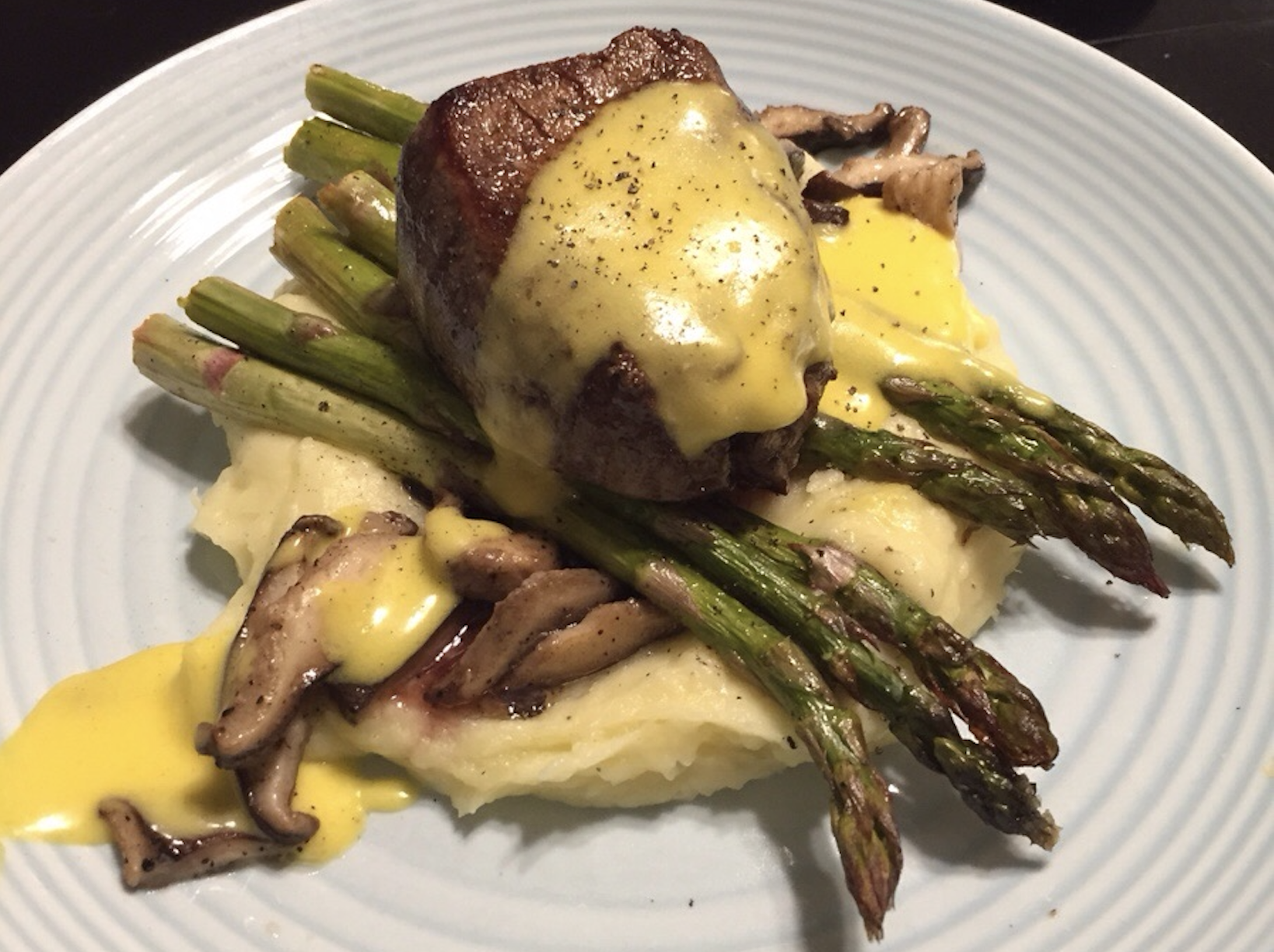 A filet mignon topped with hollandaise sauce, served on mashed potatoes with grilled asparagus and sautéed mushrooms on a white plate.