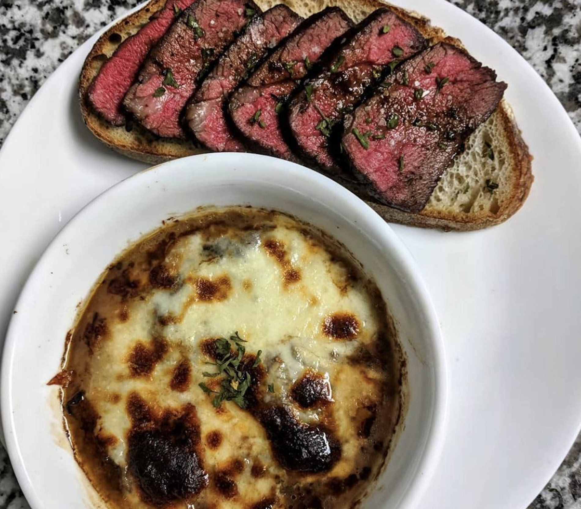 Slices of rare steak on toasted bread beside a bowl of cheesy, browned French onion soup on a white plate.