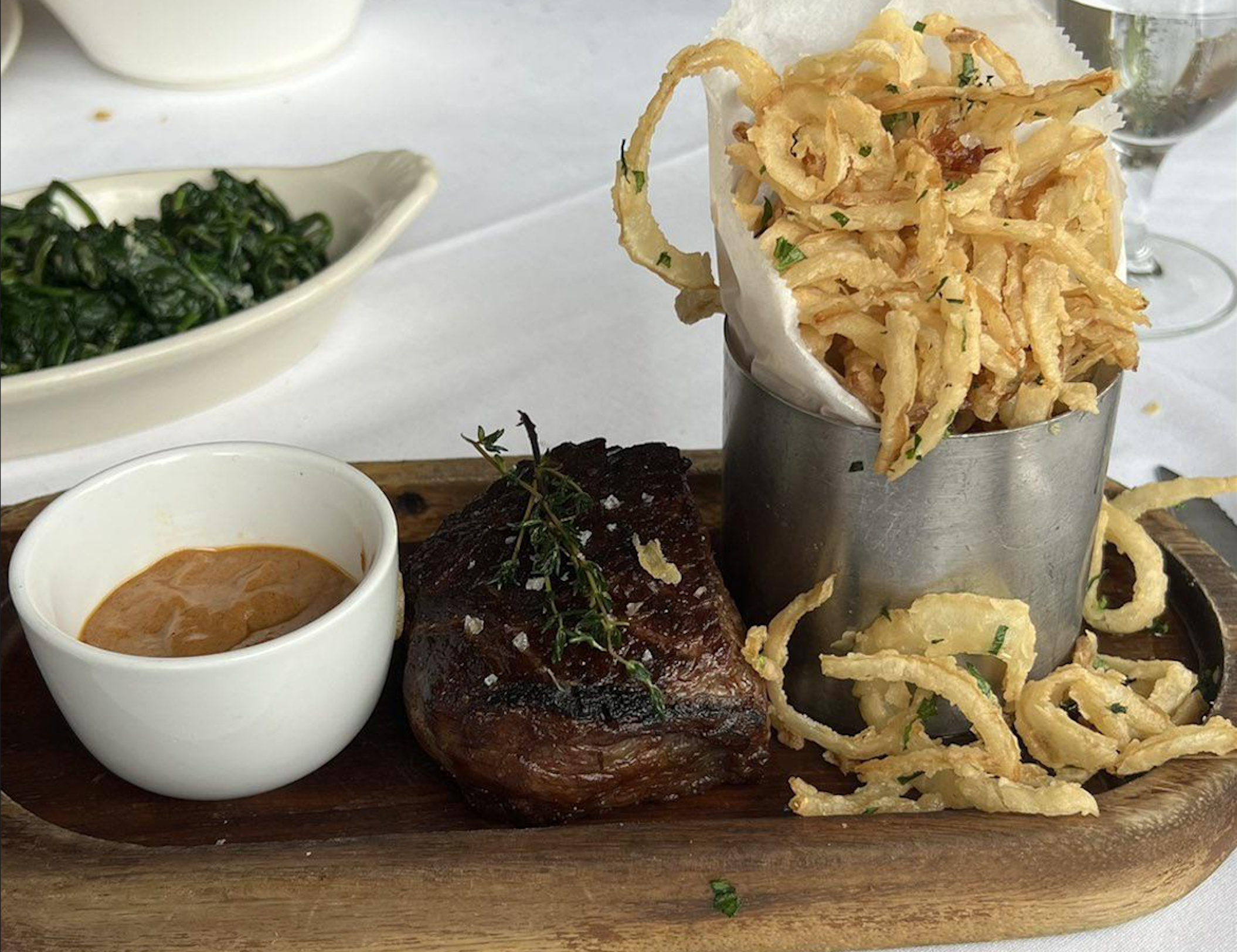 A wooden board with grilled steak topped with herbs, a cup of sauce, crispy onion strings in a metal container, and a dish of sautéed spinach in the background.
