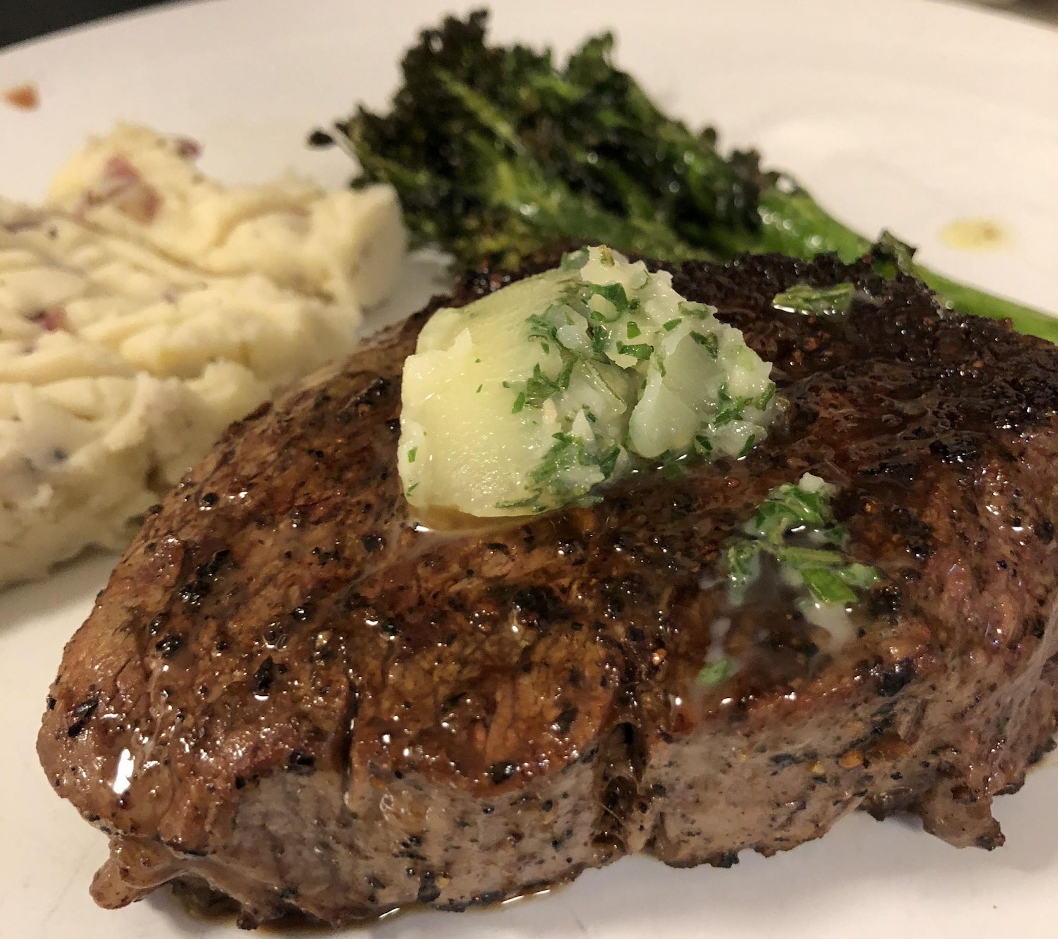 A grilled steak topped with herb butter, served with mashed potatoes and broccolini on a white plate.