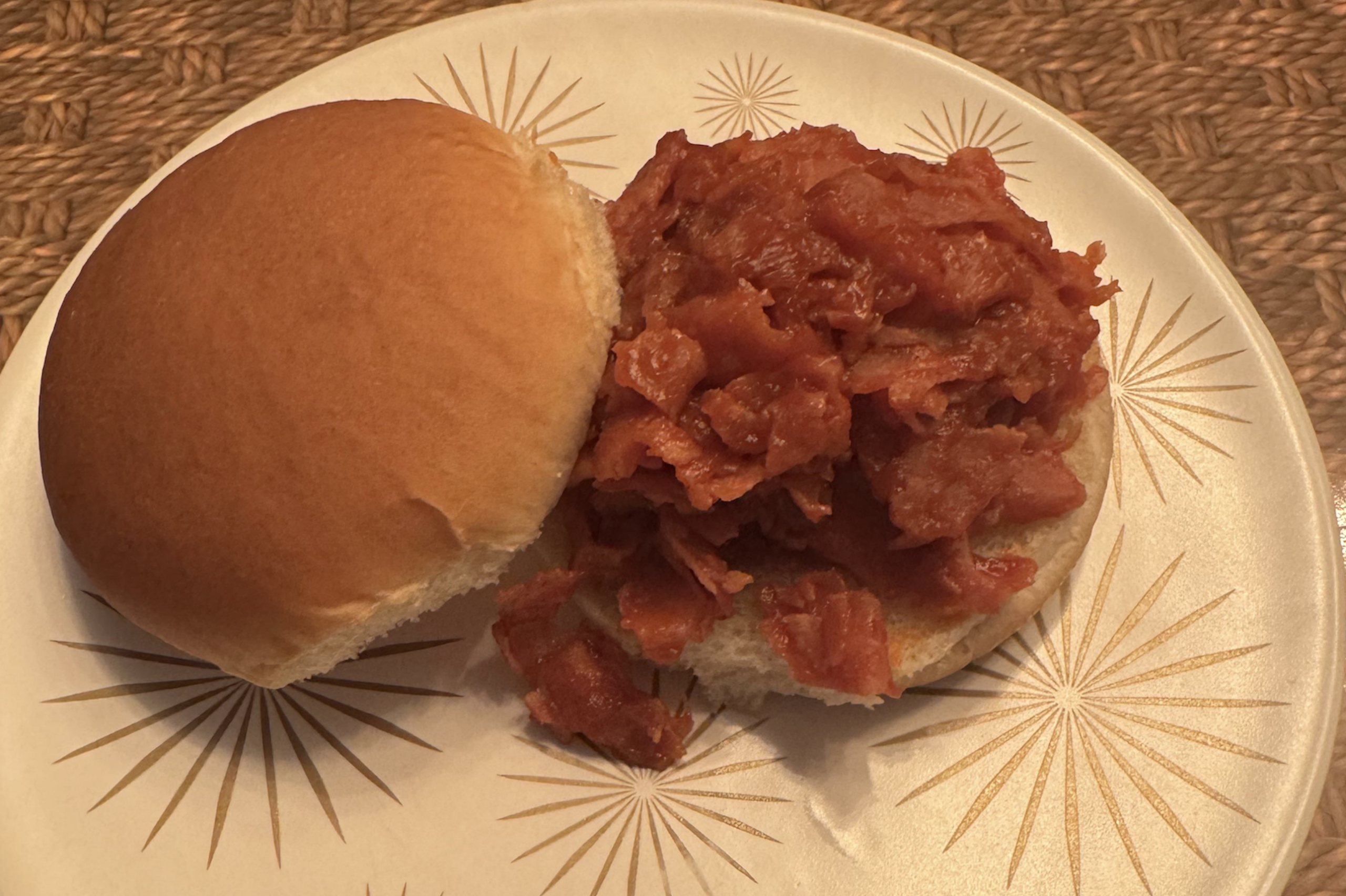 An open sandwich bun filled with a chunky tomato-red mixture, served on a white plate with gold starburst designs atop a woven placemat.