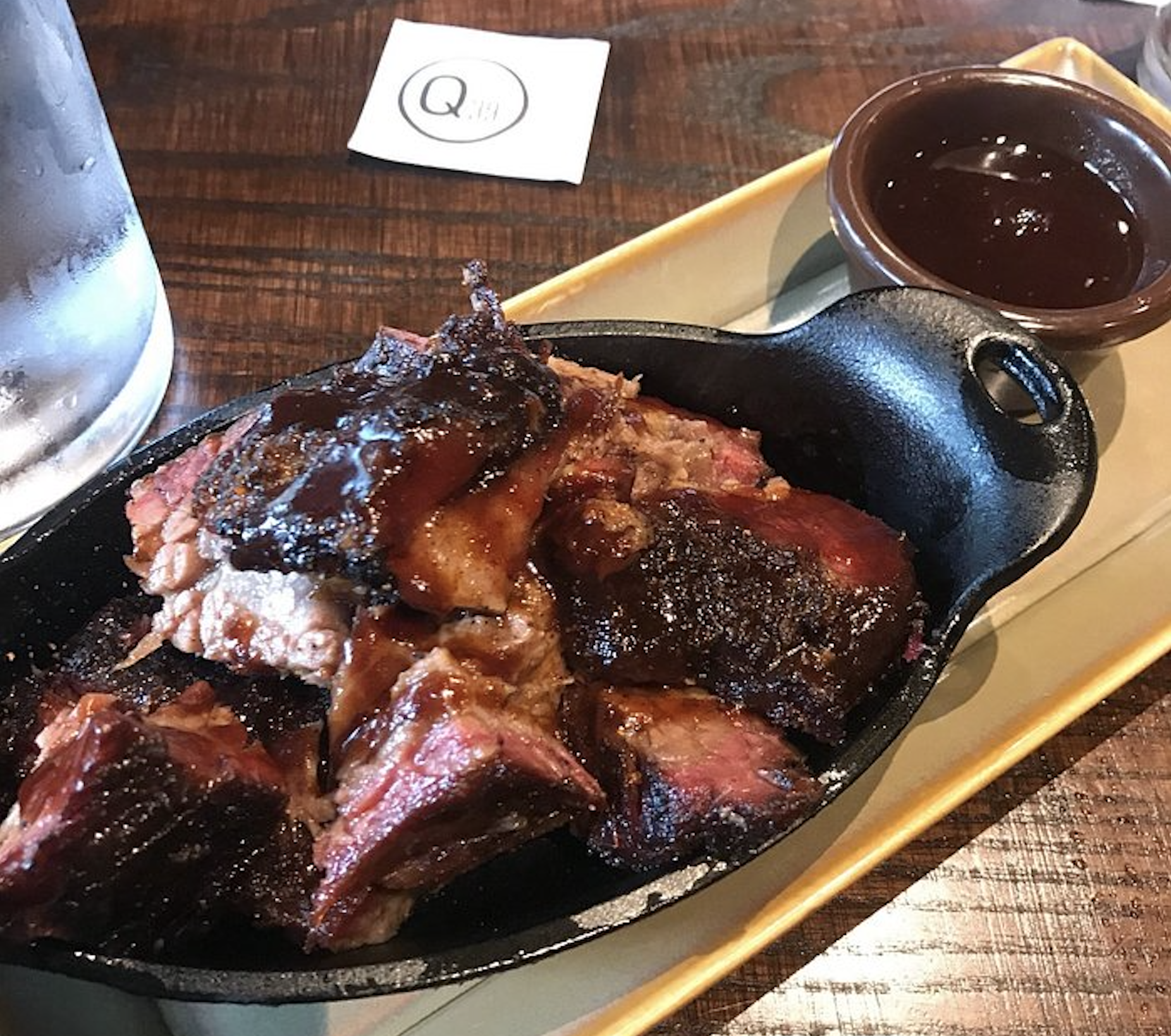 Chunks of smoked brisket burnt ends in a small black skillet, served with a side of barbecue sauce on a rectangular plate. A glass of water and a napkin are on the wooden table nearby.