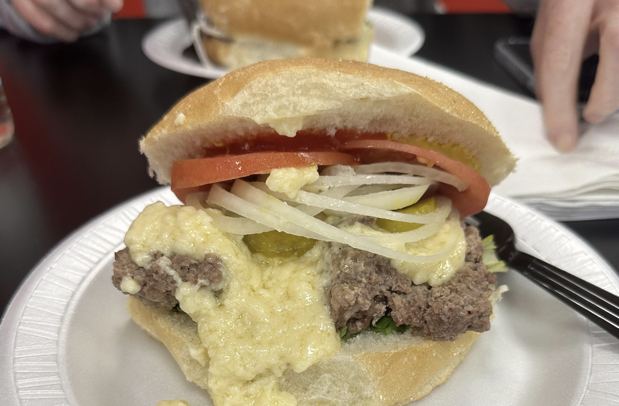 A close-up of a cheeseburger on a white plate, topped with melted cheese, onions, tomato slices, and pickles. A person’s hand is partially visible in the background.