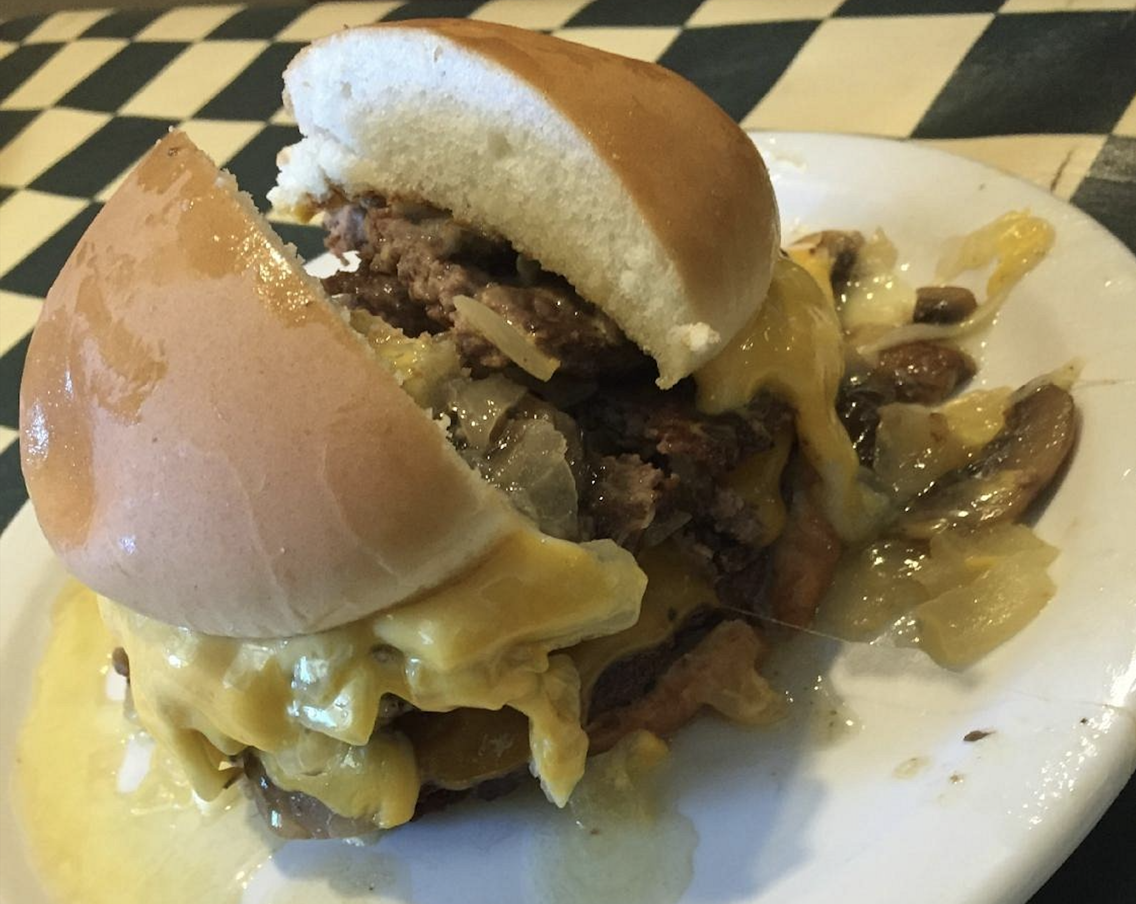 A cheeseburger with melted cheese, onions, and mushrooms overflows onto a white plate, sitting on a checkered tablecloth.