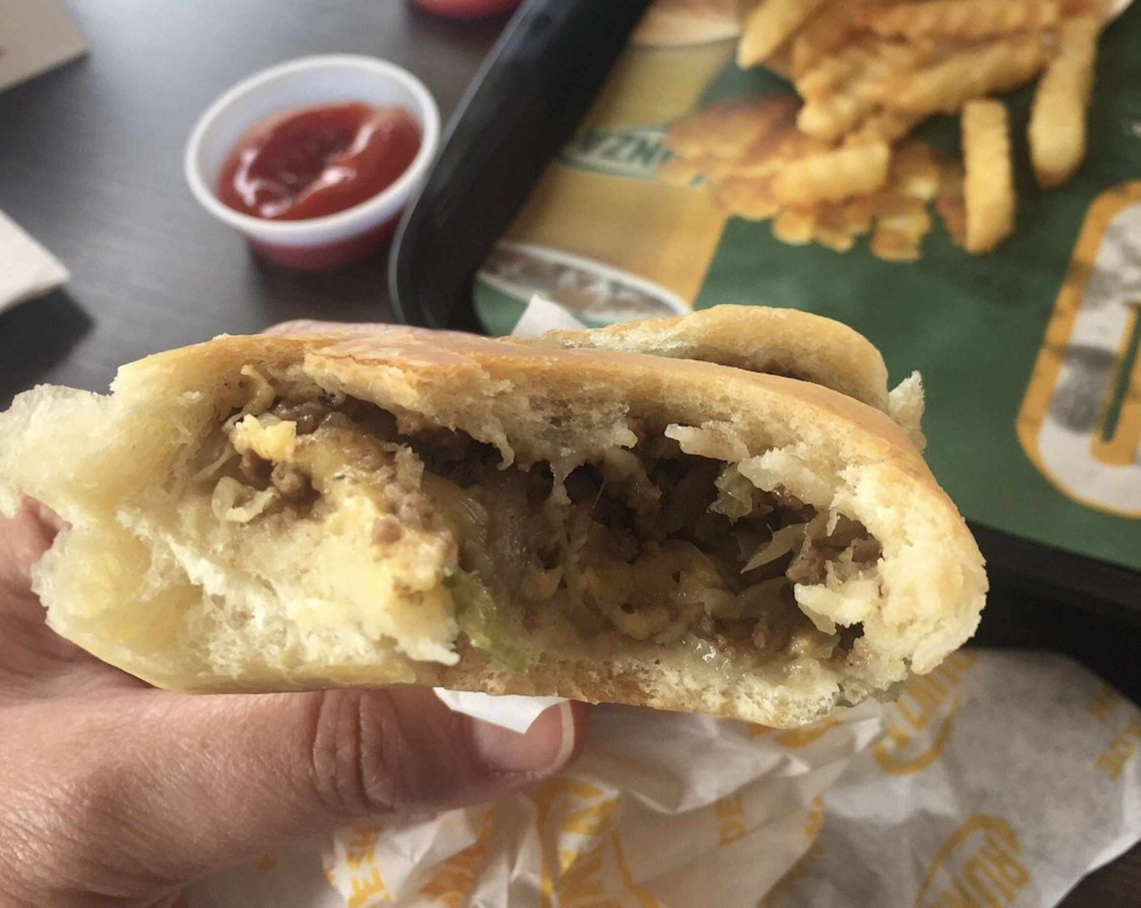 A hand holds a half-eaten stuffed bread roll showing ground meat and cheese filling, one of the American city foods. In the background, crinkle-cut fries and a cup of ketchup sit on a tray.