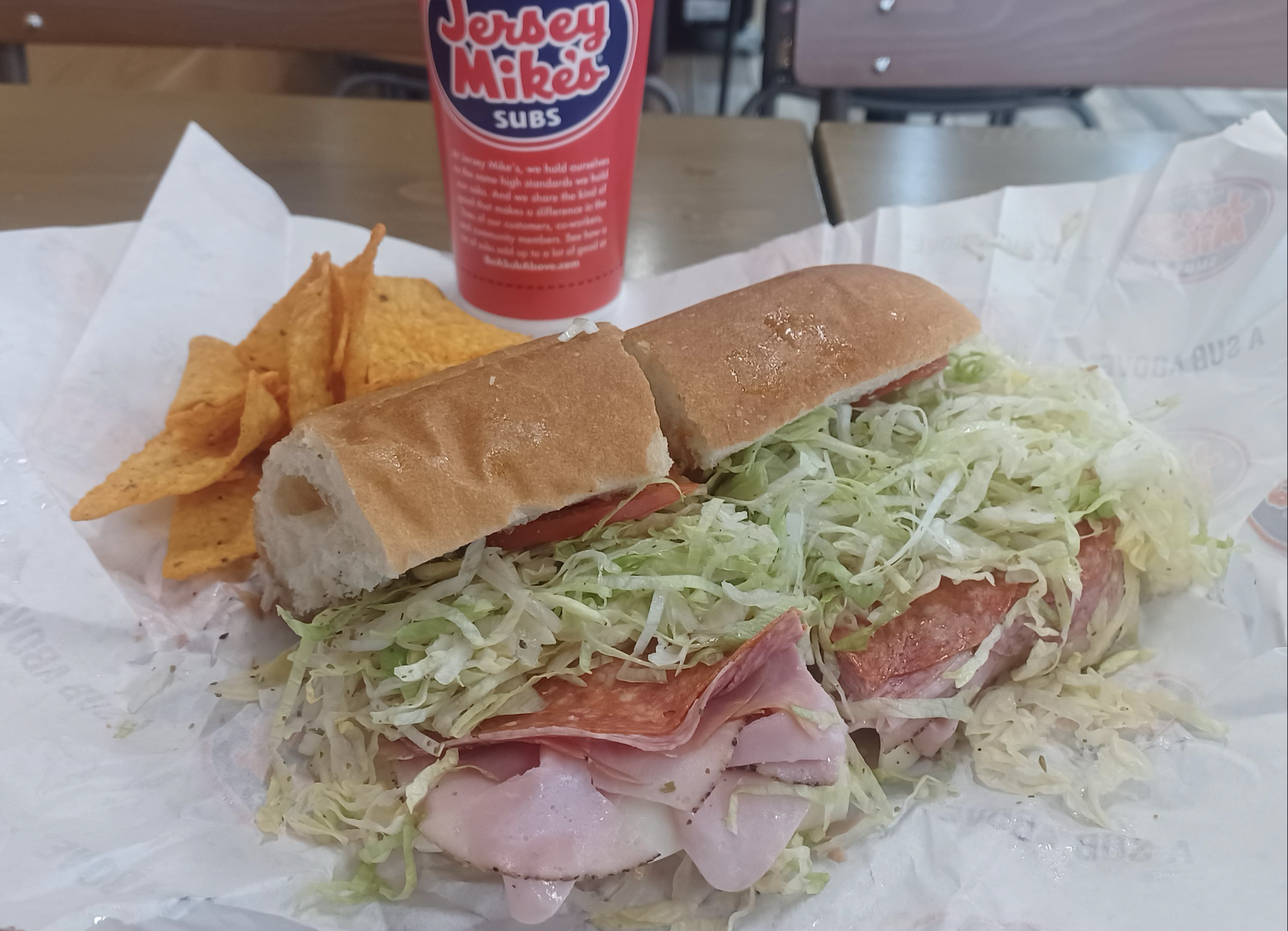 A Jersey Mike’s sub sandwich filled with ham, salami, and shredded lettuce sits on paper next to Doritos chips and a Jersey Mike’s cup on a table in a casual dining setting.