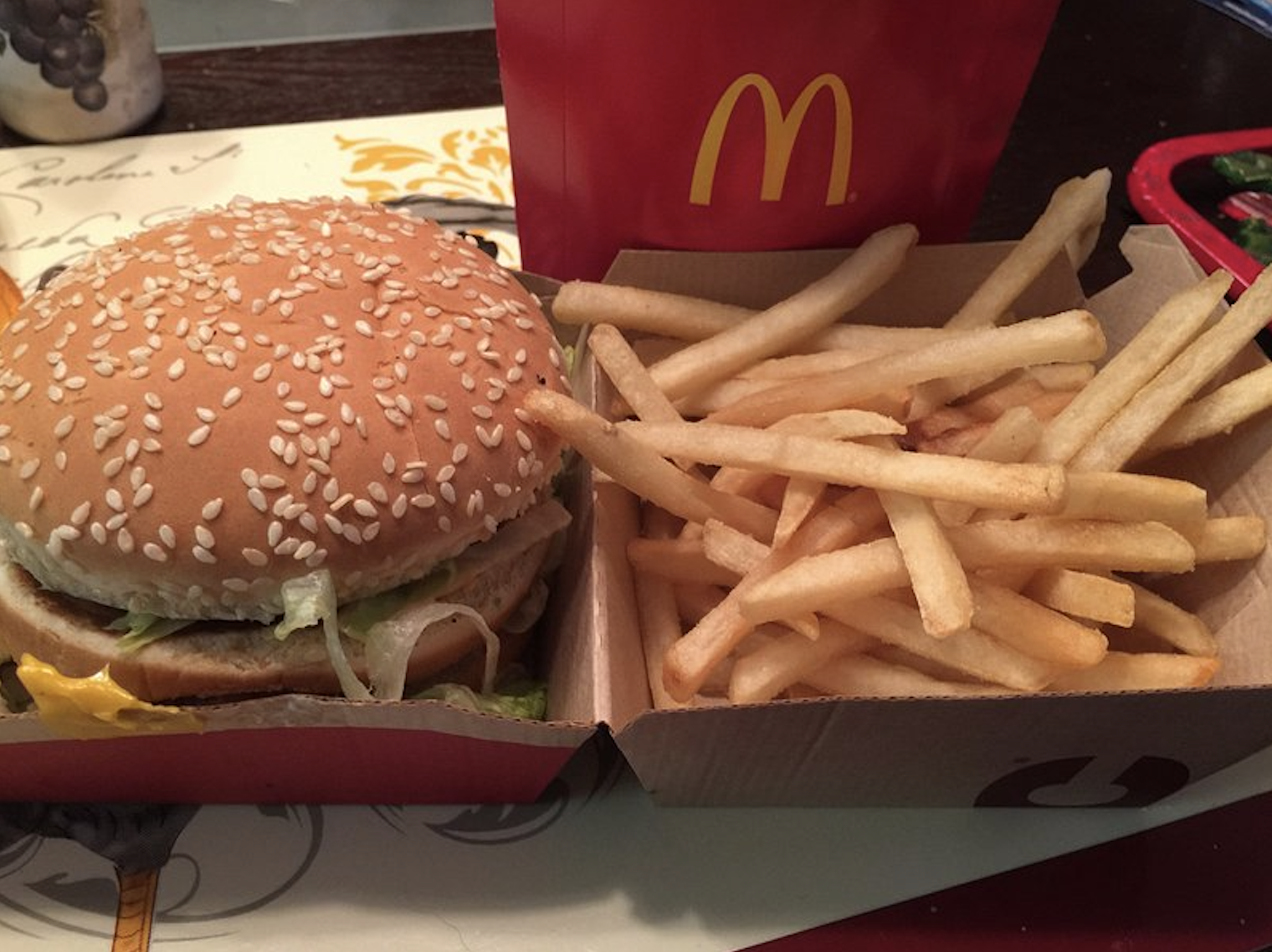 A McDonald’s meal with a sesame seed burger and a serving of French fries in a cardboard box, with a red McDonald’s fries container in the background.