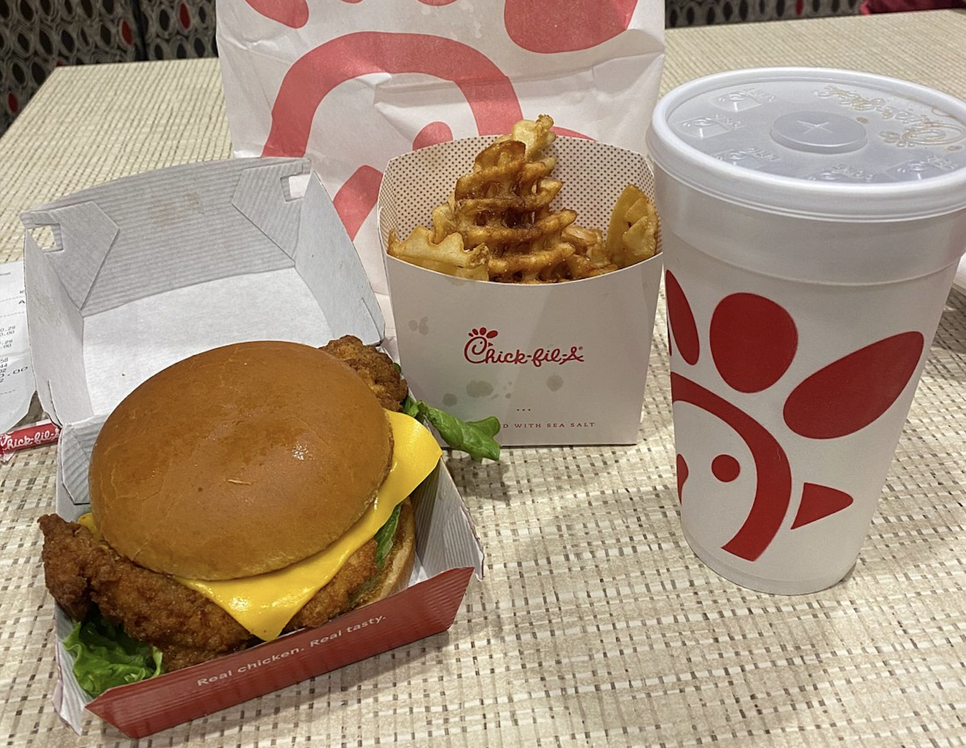 A Chick-fil-A meal with a chicken sandwich topped with cheese, waffle fries in a branded container, and a large drink cup, all on a woven table surface.