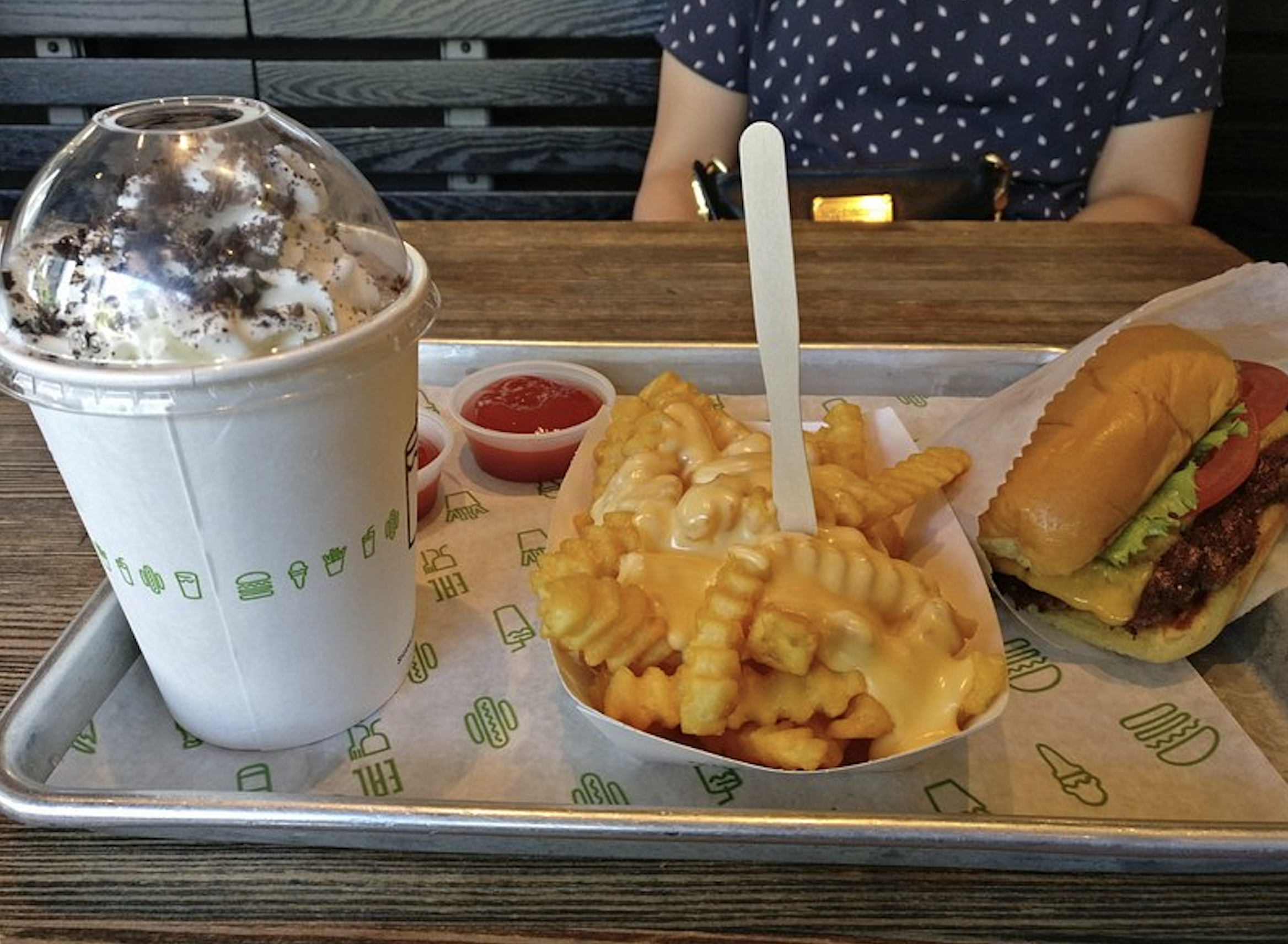 A tray with crinkle-cut fries topped with cheese, a cheeseburger with lettuce and tomato, ketchup cups, and a large milkshake with whipped cream and cookie bits. A person in a polka dot shirt sits behind the tray.