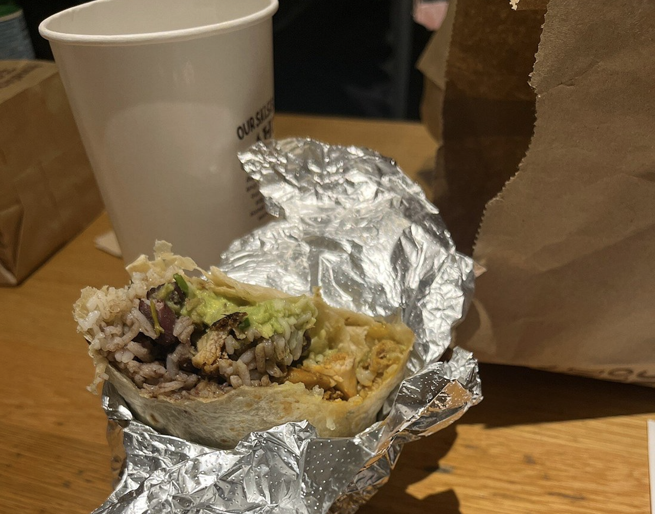 A partially eaten burrito wrapped in foil sits on a wooden table next to a white paper cup and a brown paper bag.