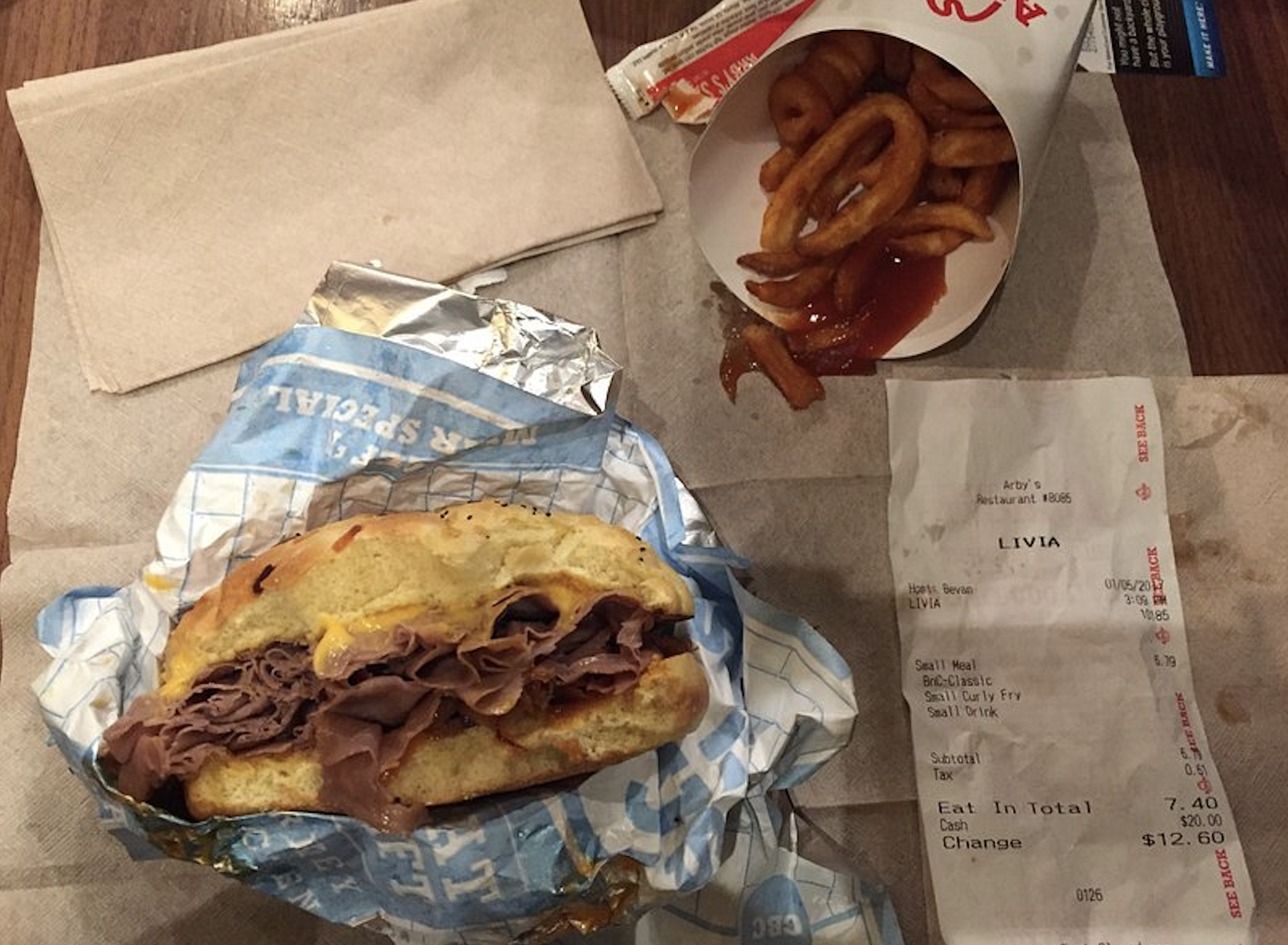 A roast beef sandwich sits on a wrapper next to curly fries with ketchup in a paper container, a ketchup packet, and a receipt, all on a brown table.