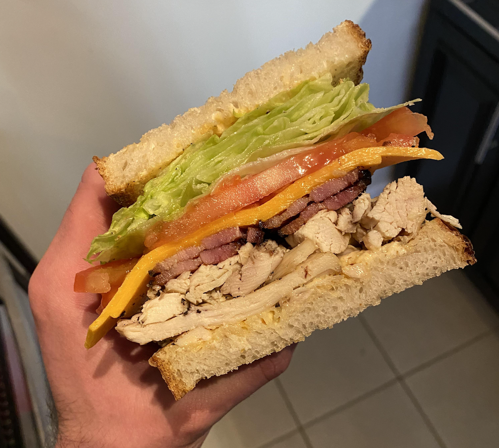 A hand holds a thick sandwich filled with lettuce, tomato, cheddar cheese, bacon, and sliced chicken between two slices of white bread.