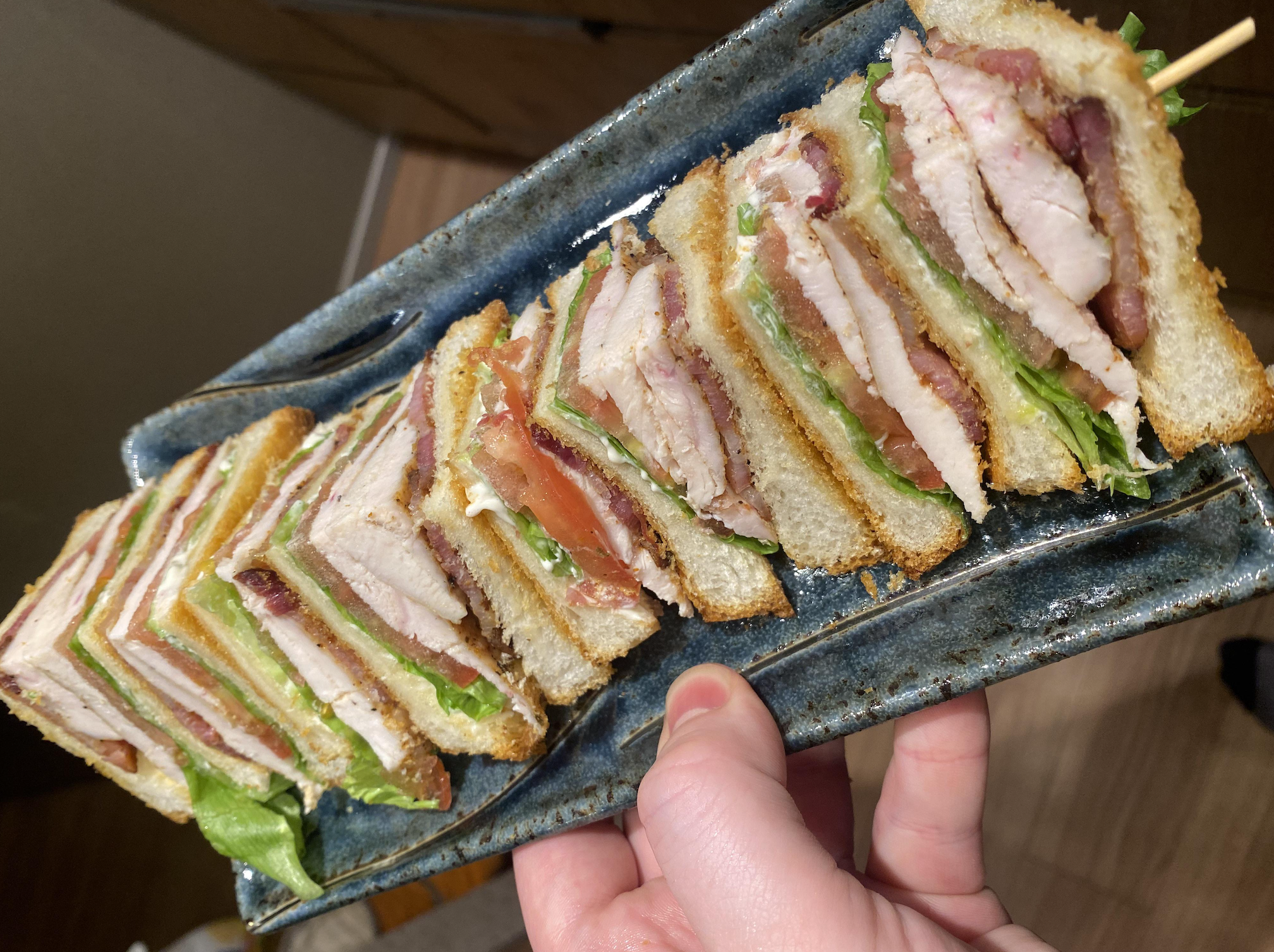 A hand holds a rectangular plate with neatly arranged club sandwiches filled with layers of meat, lettuce, and tomato on sliced bread.
