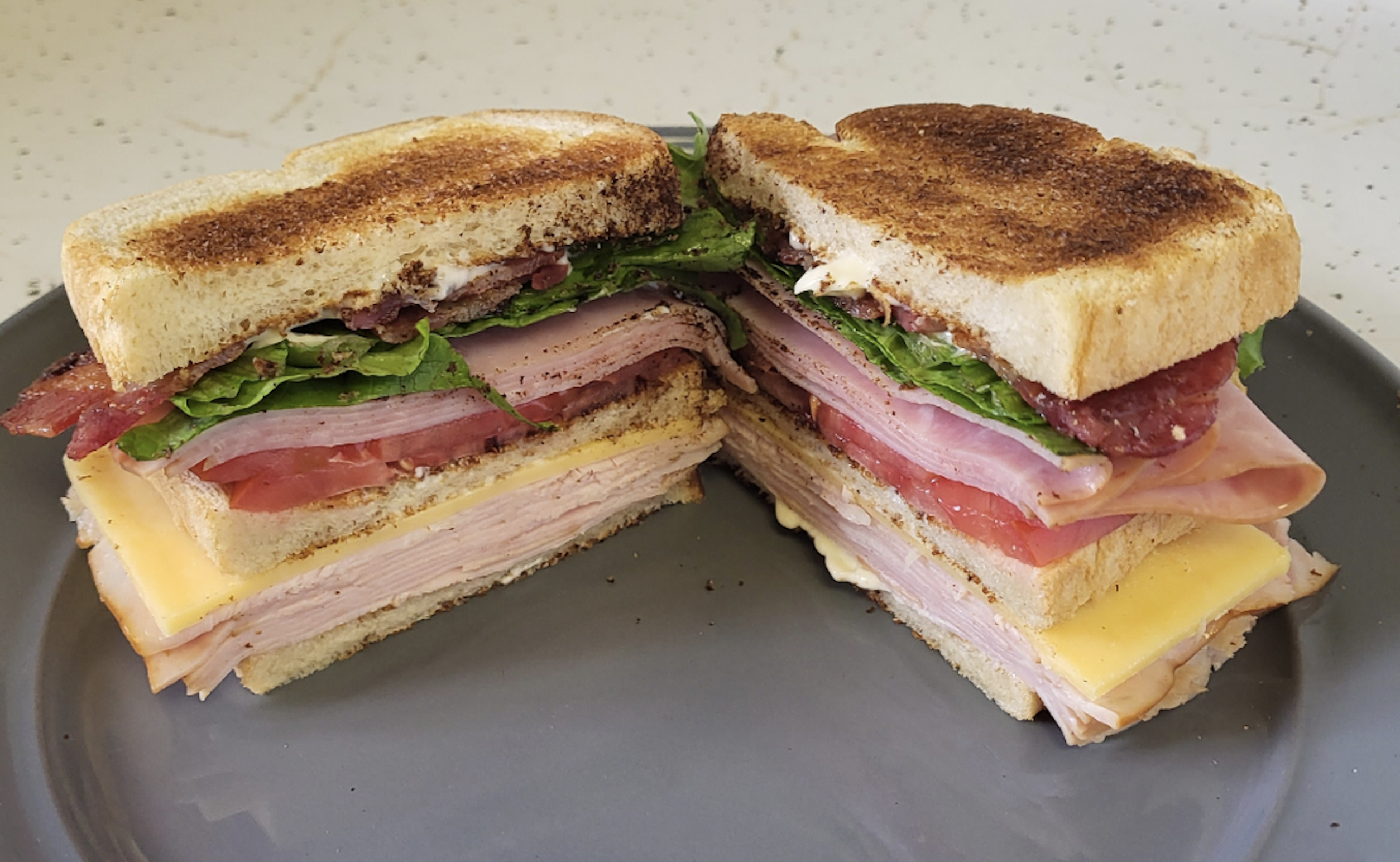 A club sandwich with toasted bread, layers of turkey, ham, cheddar cheese, lettuce, tomato, and bacon, cut in half and served on a gray plate.