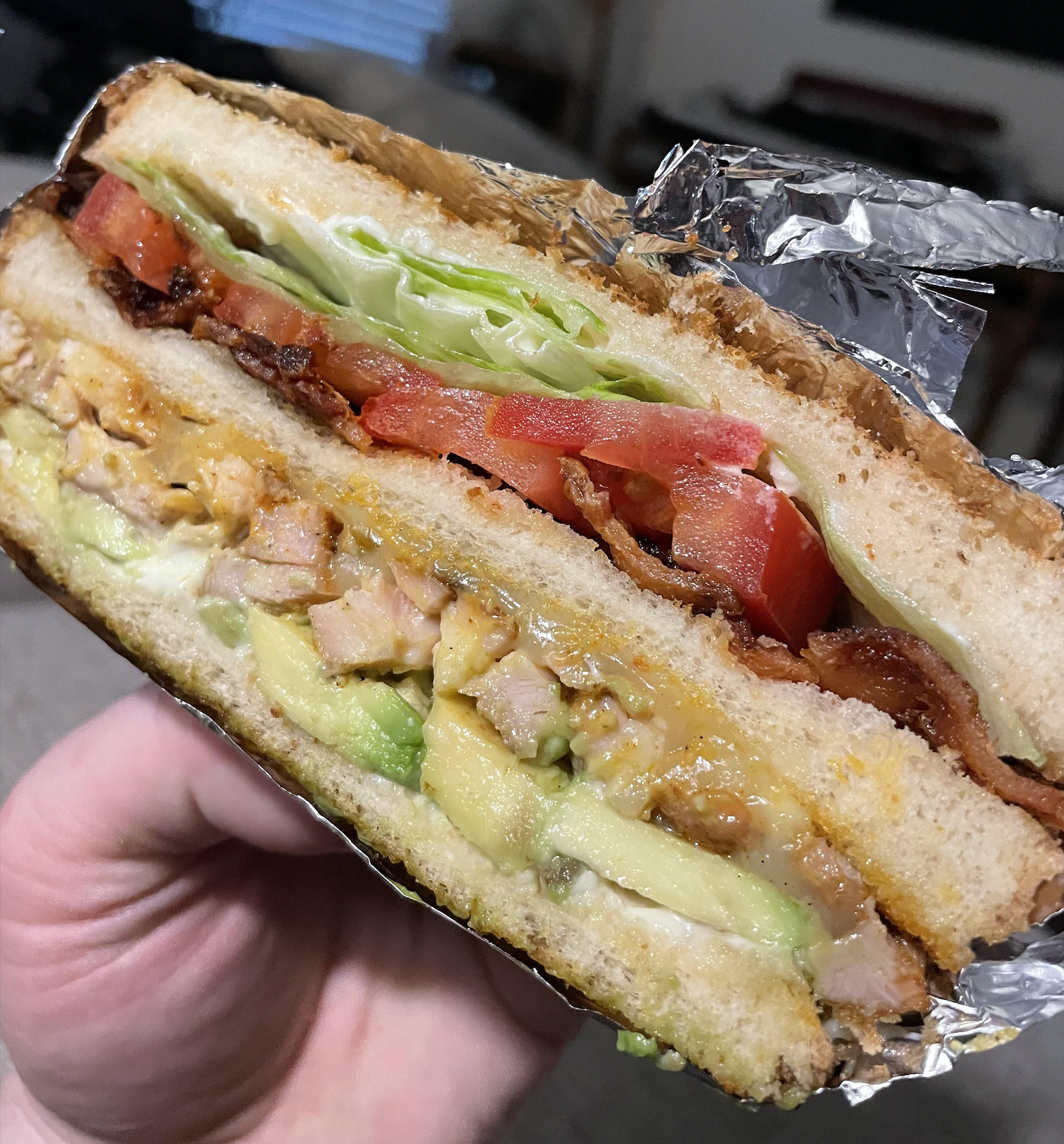 A hand holds a thick sandwich with toasted bread, lettuce, tomato, bacon, avocado, melted cheese, and grilled chicken, wrapped partly in aluminum foil.