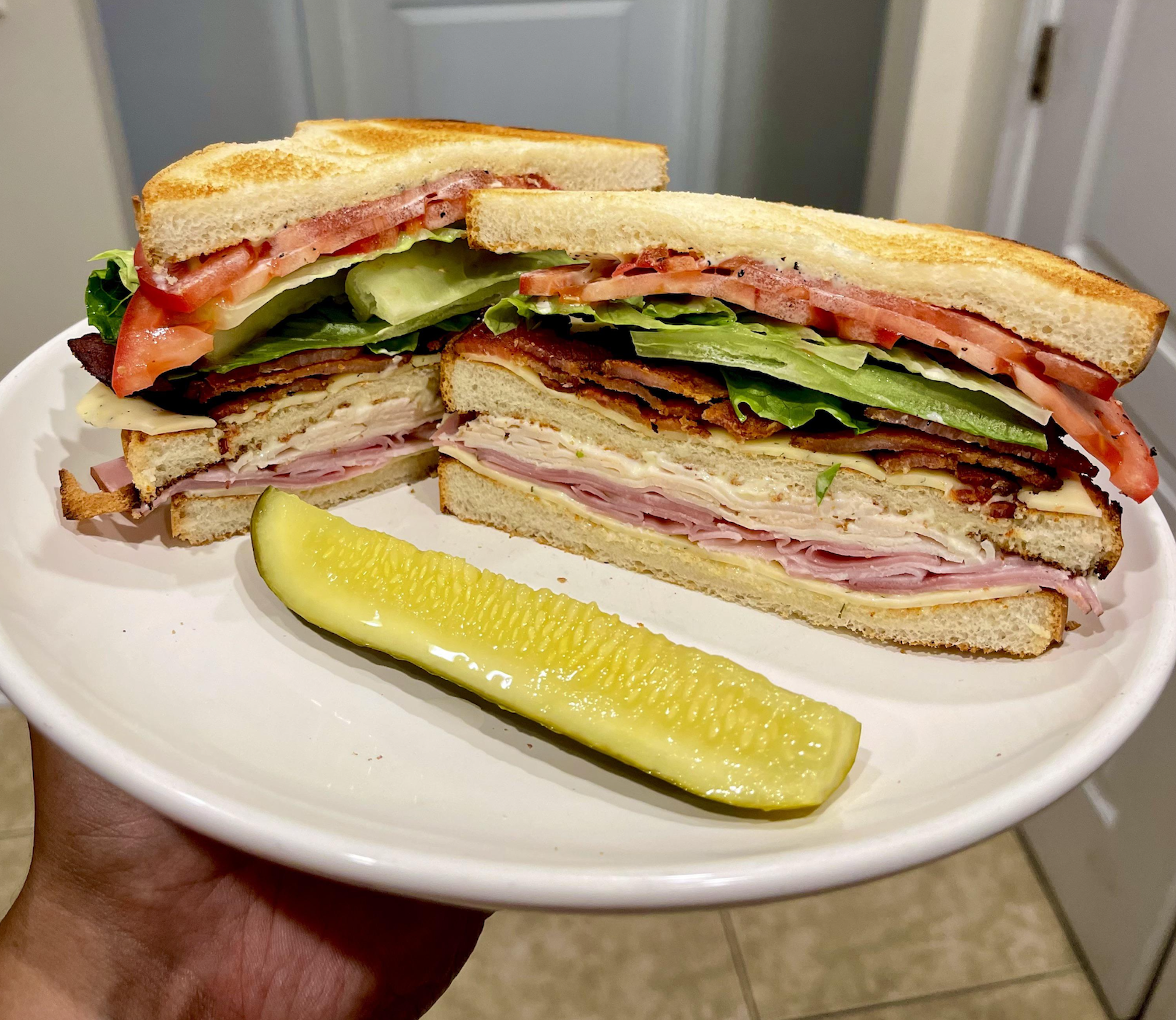 A plate holds a club sandwich cut in half, showing layers of ham, turkey, bacon, lettuce, tomato, and cucumber on toasted bread, next to a dill pickle spear.