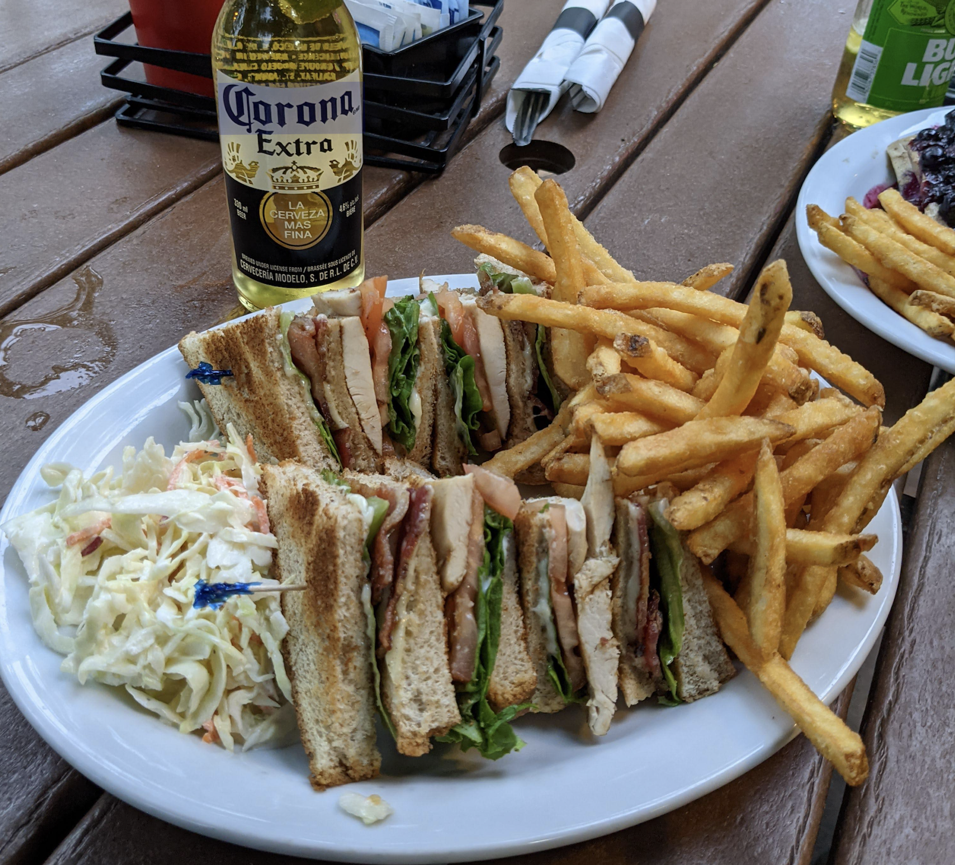 A plate with a club sandwich cut in quarters, French fries, and coleslaw sits on a wooden table next to a bottle of Corona Extra beer.
