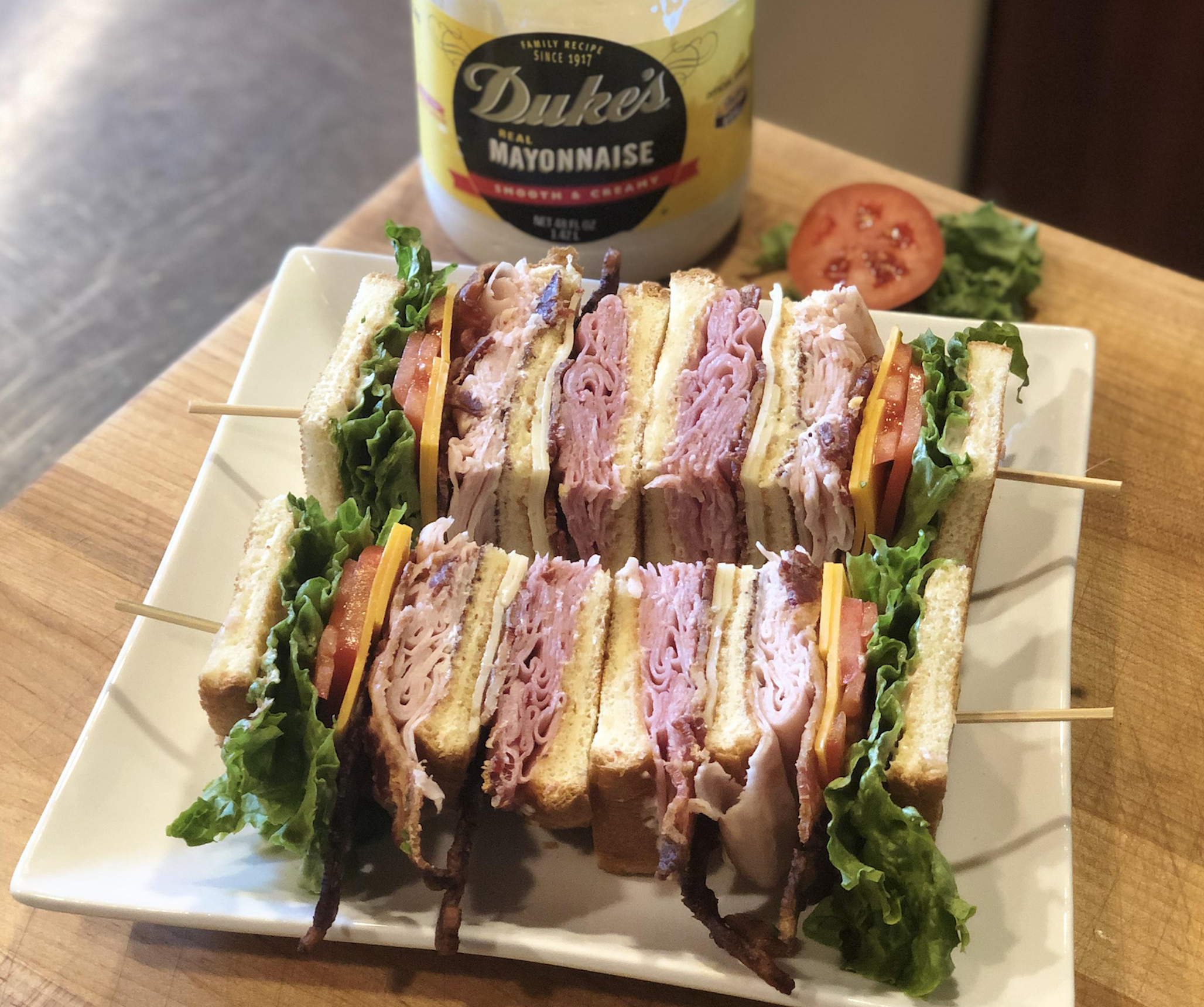 A plate of club sandwiches with layers of sliced turkey, ham, lettuce, tomato, and bacon, secured with wooden picks. A jar of Duke’s mayonnaise, lettuce, and a tomato slice are in the background.