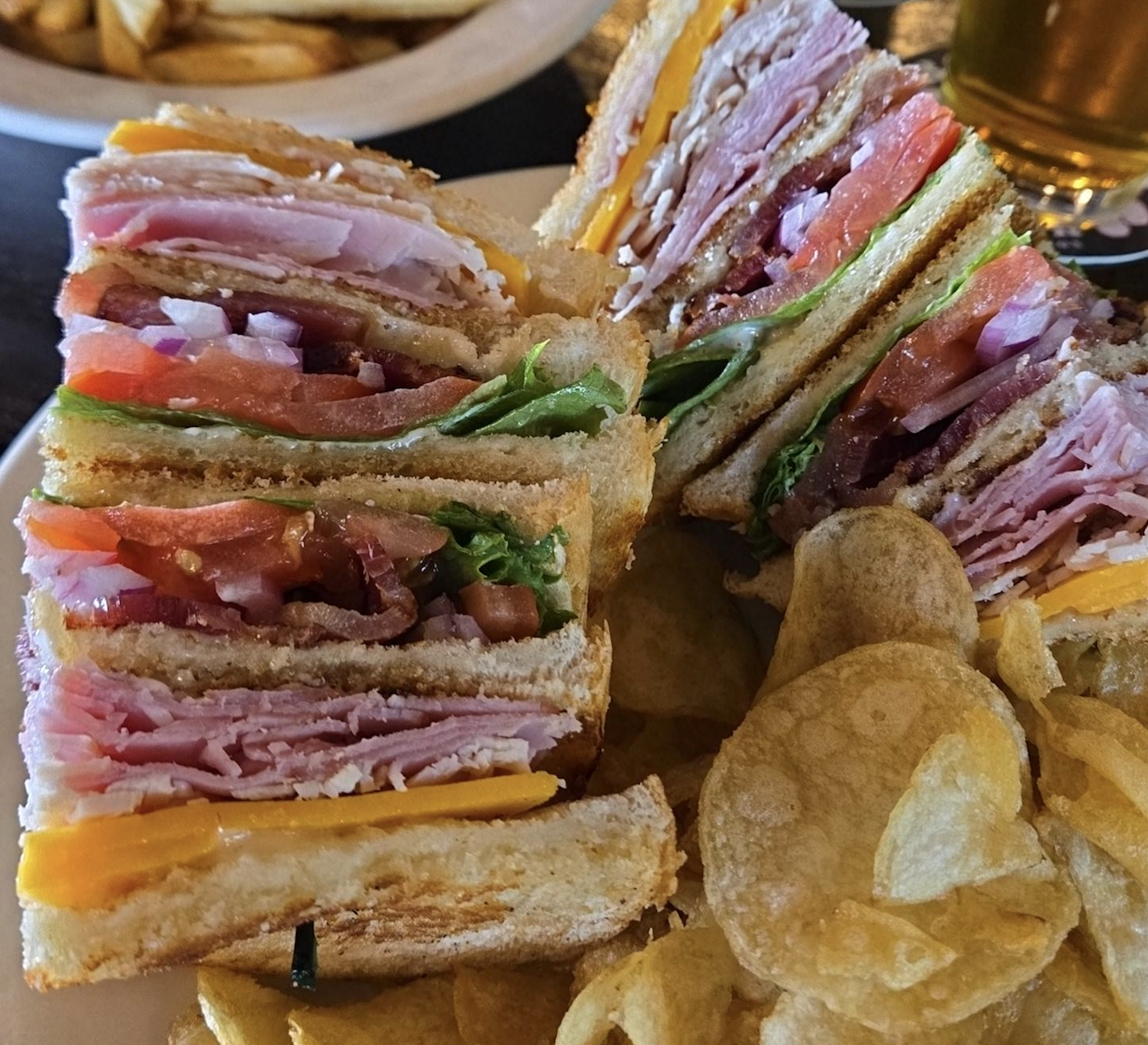 A close-up of a club sandwich with layers of ham, turkey, cheddar cheese, lettuce, tomato, and bacon, served with potato chips on a white plate.