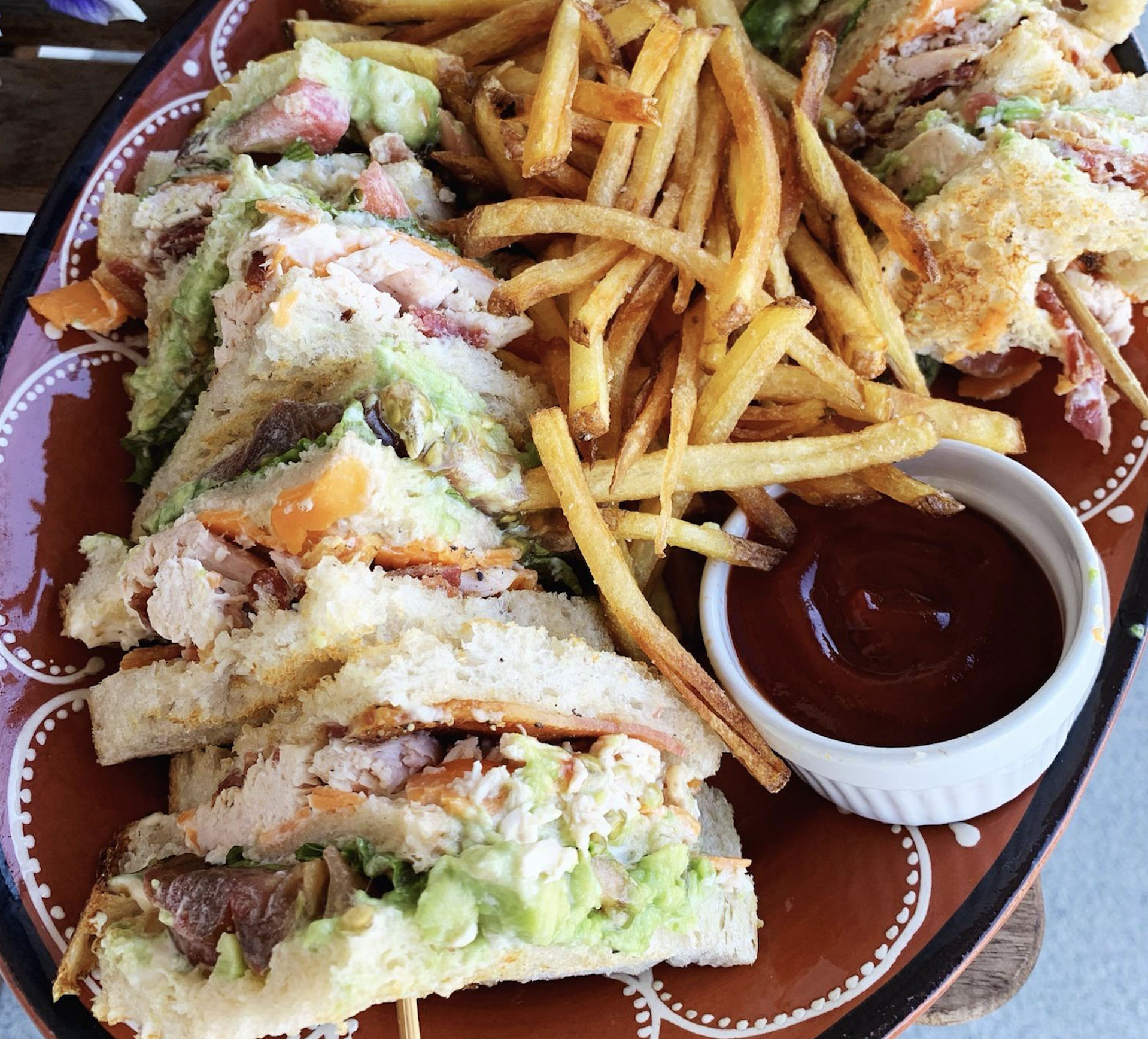 A plate of club sandwiches filled with chicken, lettuce, tomato, and avocado, served with thin french fries and a small cup of ketchup.
