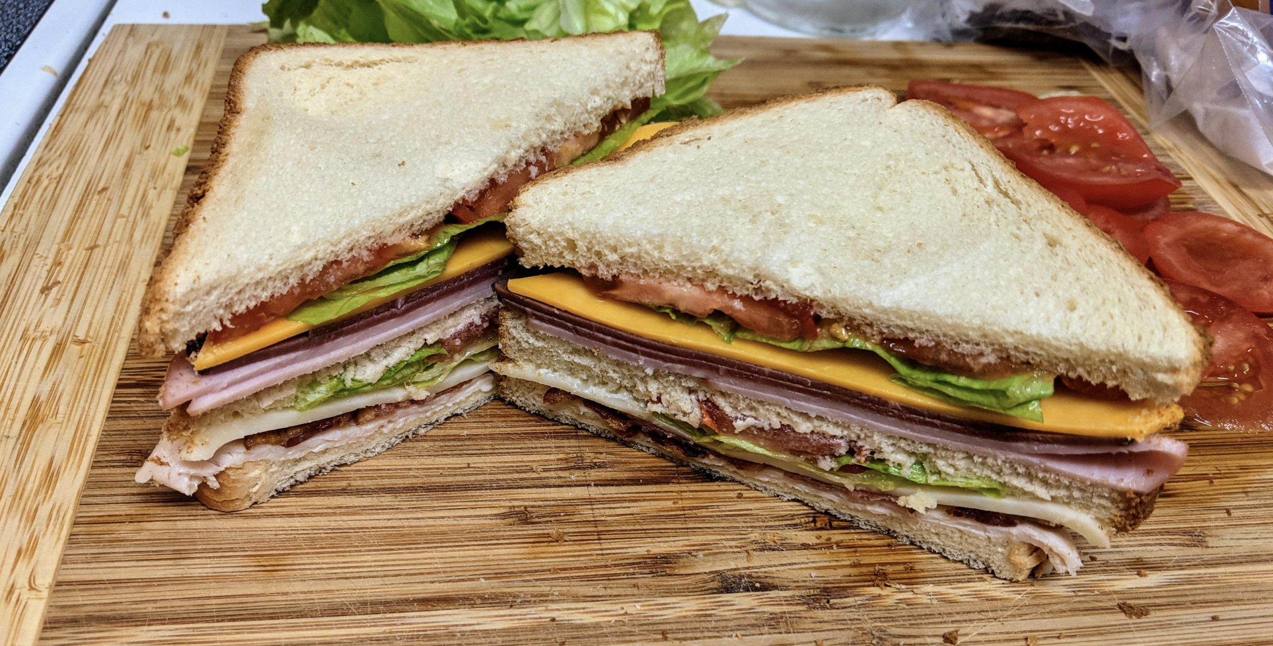 A sandwich cut in half on a wooden board, layered with lettuce, tomato, cheddar cheese, ham, turkey, and white bread. Slices of tomato and lettuce leaves are in the background.