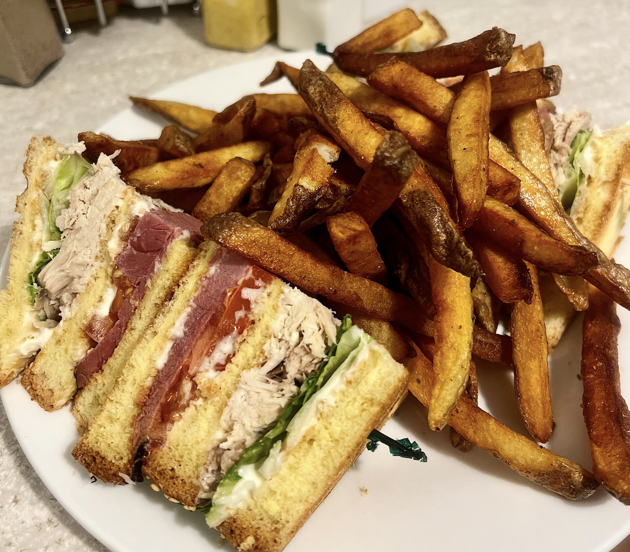 A plate of golden French fries next to a sliced club sandwich filled with turkey, bacon, lettuce, tomato, and mayonnaise on toasted bread.