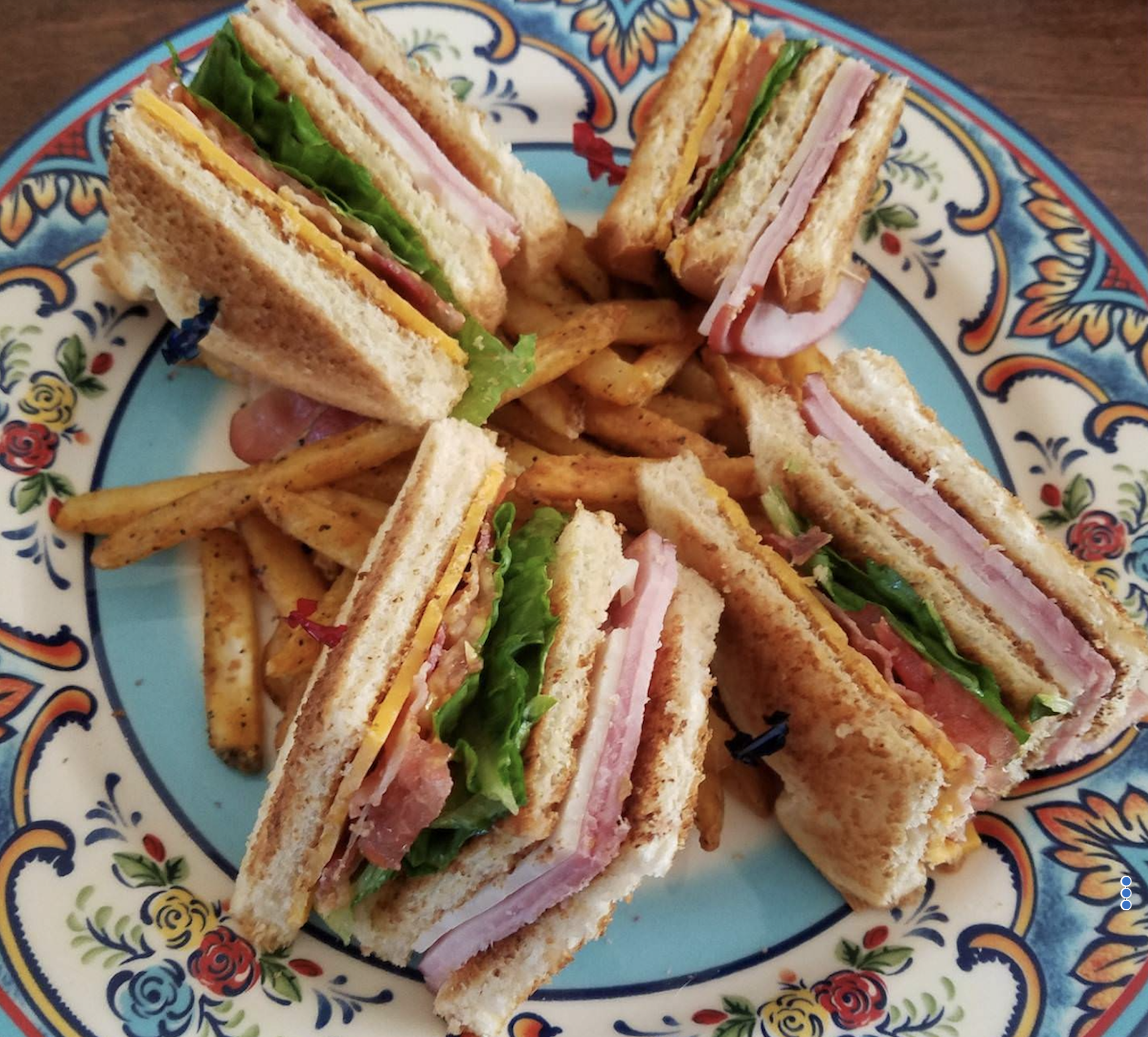 Five club sandwich quarters with layers of ham, turkey, cheese, lettuce, and tomato are arranged on a colorful plate with seasoned French fries in the center.