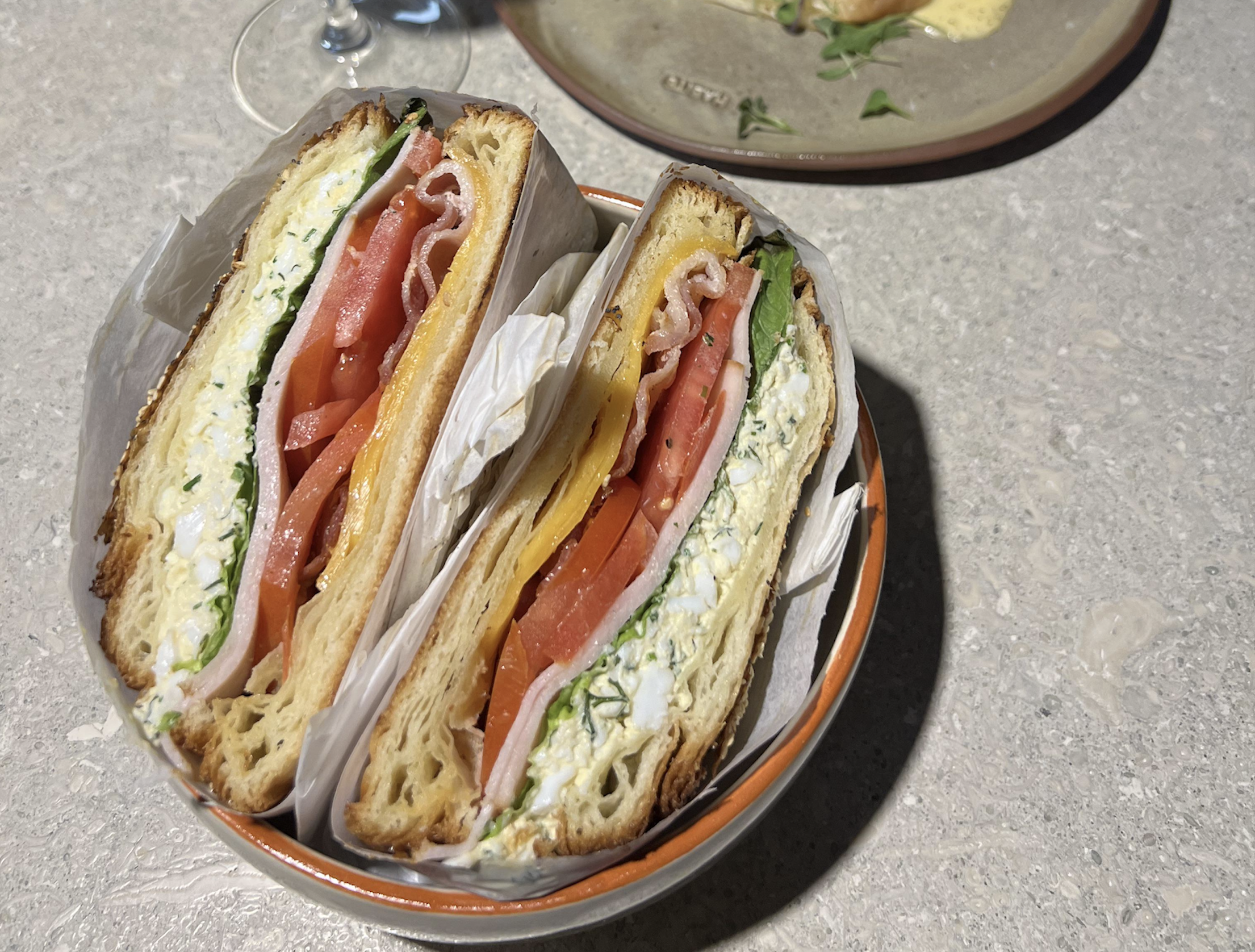 Two thick sandwiches in a bowl, filled with egg salad, tomato slices, leafy greens, and possibly ham, served on a light textured surface. A plate with food is partially visible in the background.