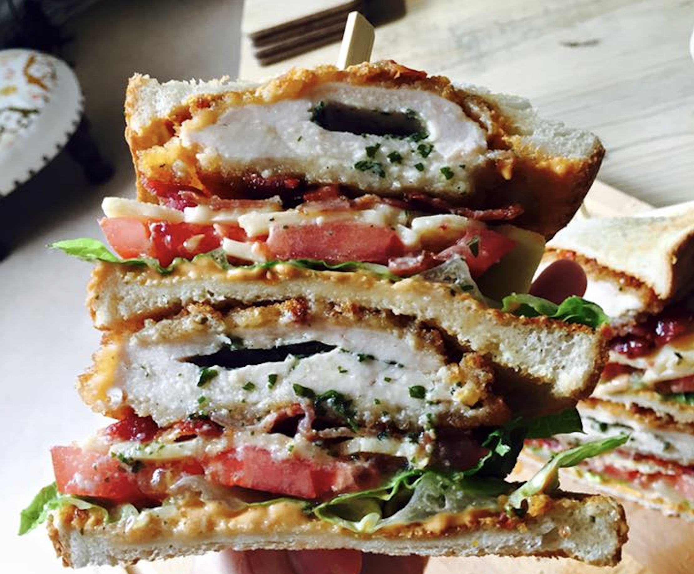 A close-up of a hand holding a stacked sandwich with crispy chicken, bacon, lettuce, tomato, and sauce between toasted bread slices.