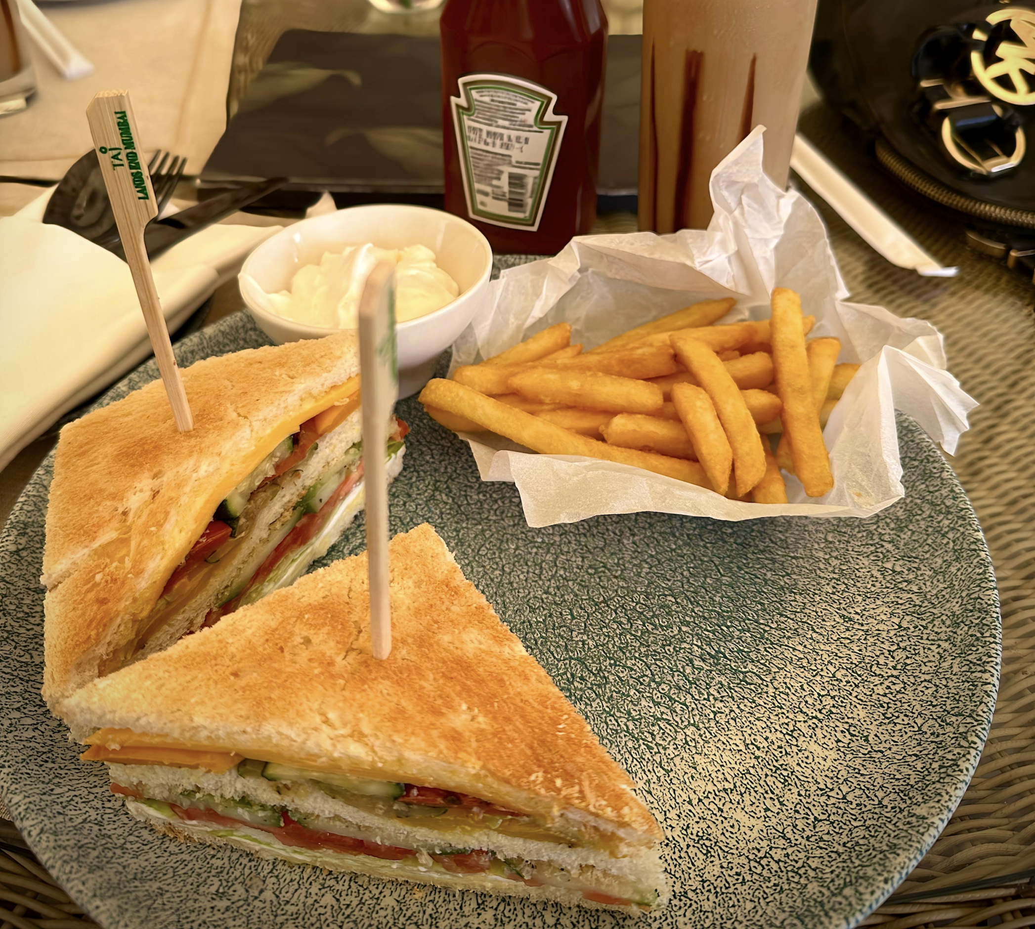 A plate with two sandwich halves, a serving of French fries in paper, a small bowl of mayonnaise, a bottle of ketchup, and a drink in the background on an outdoor table.