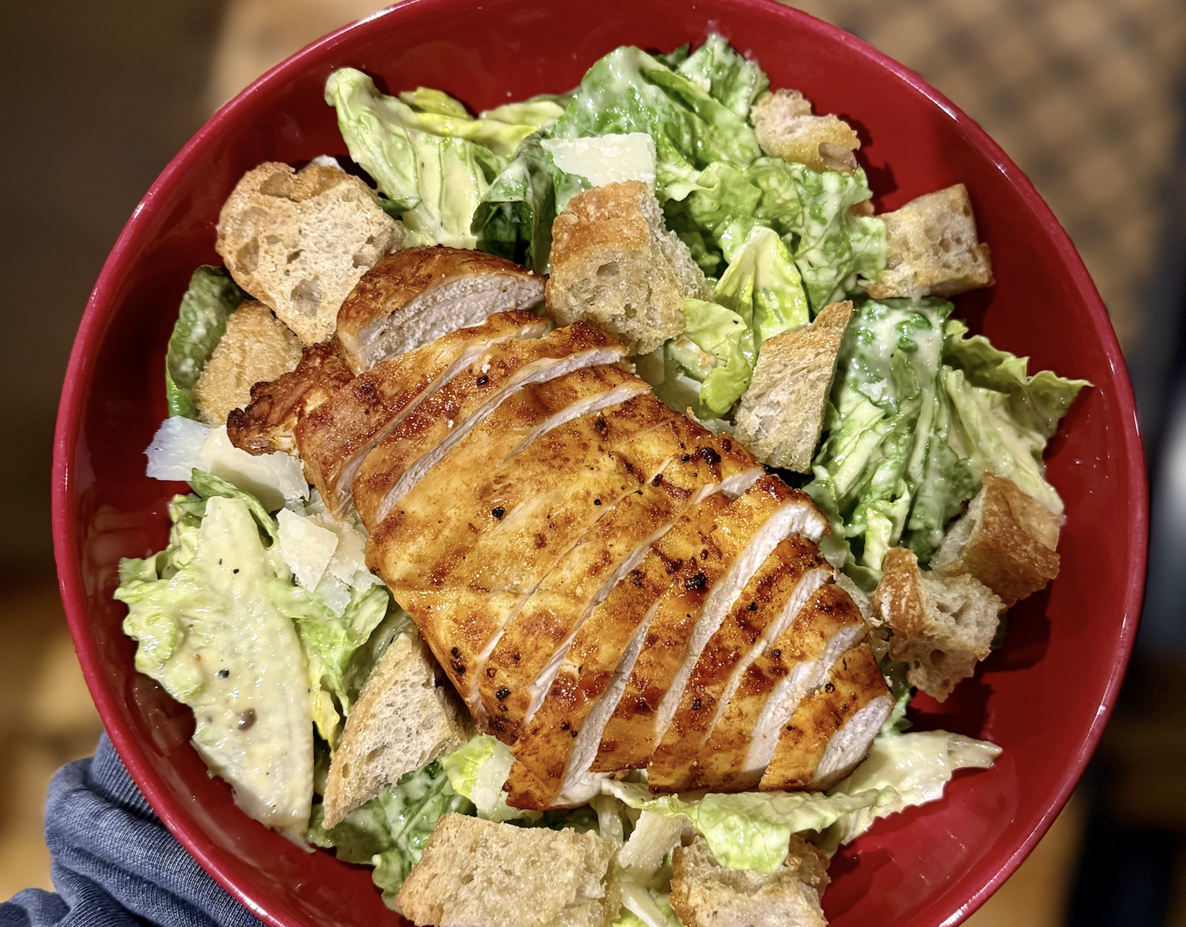 A red bowl filled with Caesar salad, topped with sliced grilled chicken breast, croutons, avocado, and romaine lettuce.