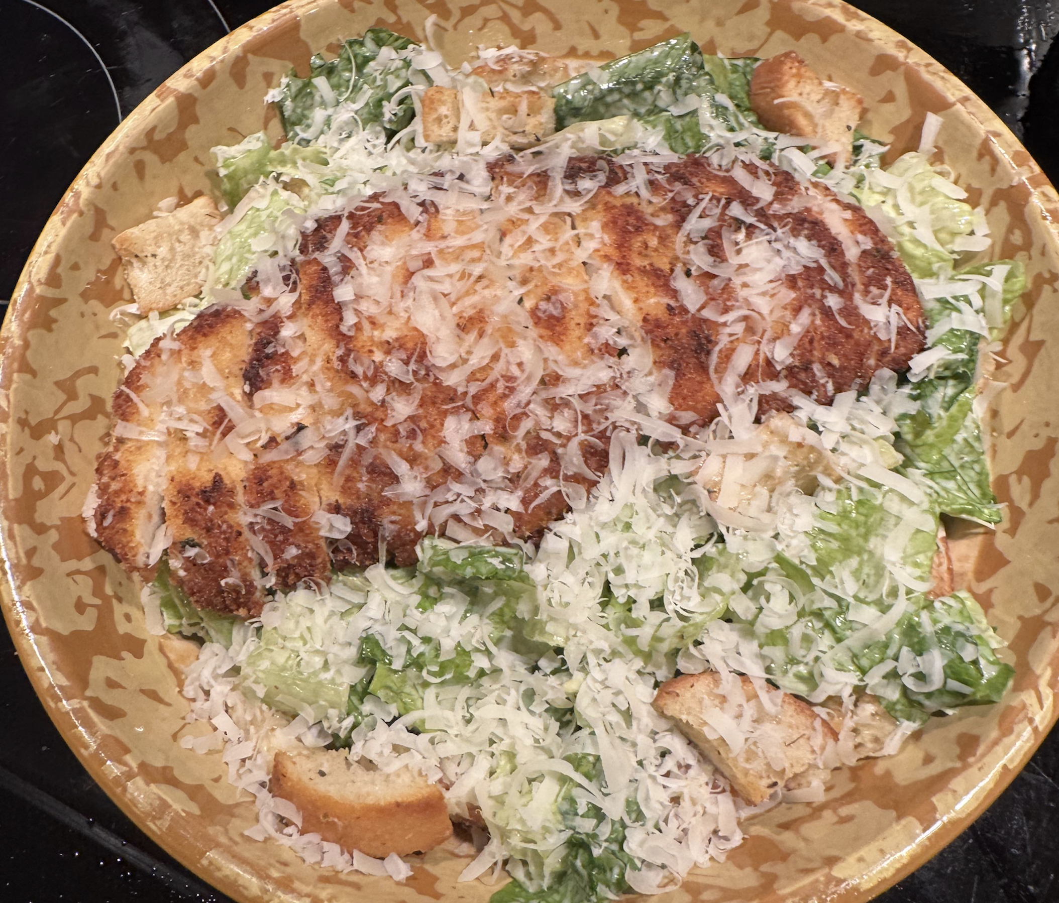 A bowl of Caesar salad topped with breaded, sliced chicken cutlet, grated cheese, romaine lettuce, and croutons.