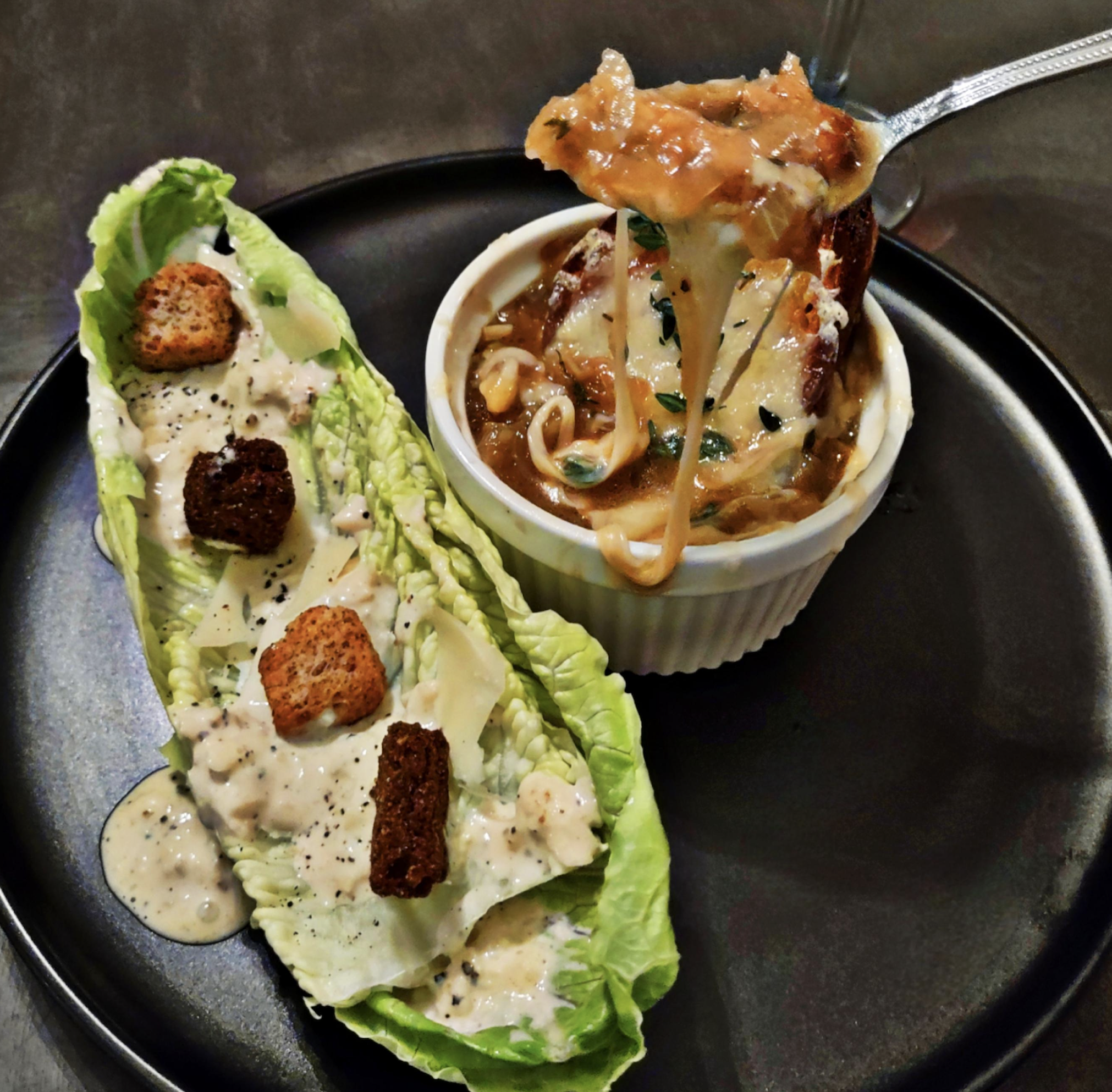 A black plate with Caesar salad topped with croutons and creamy dressing, next to a ramekin of French onion soup with melted cheese, being lifted by a spoon.