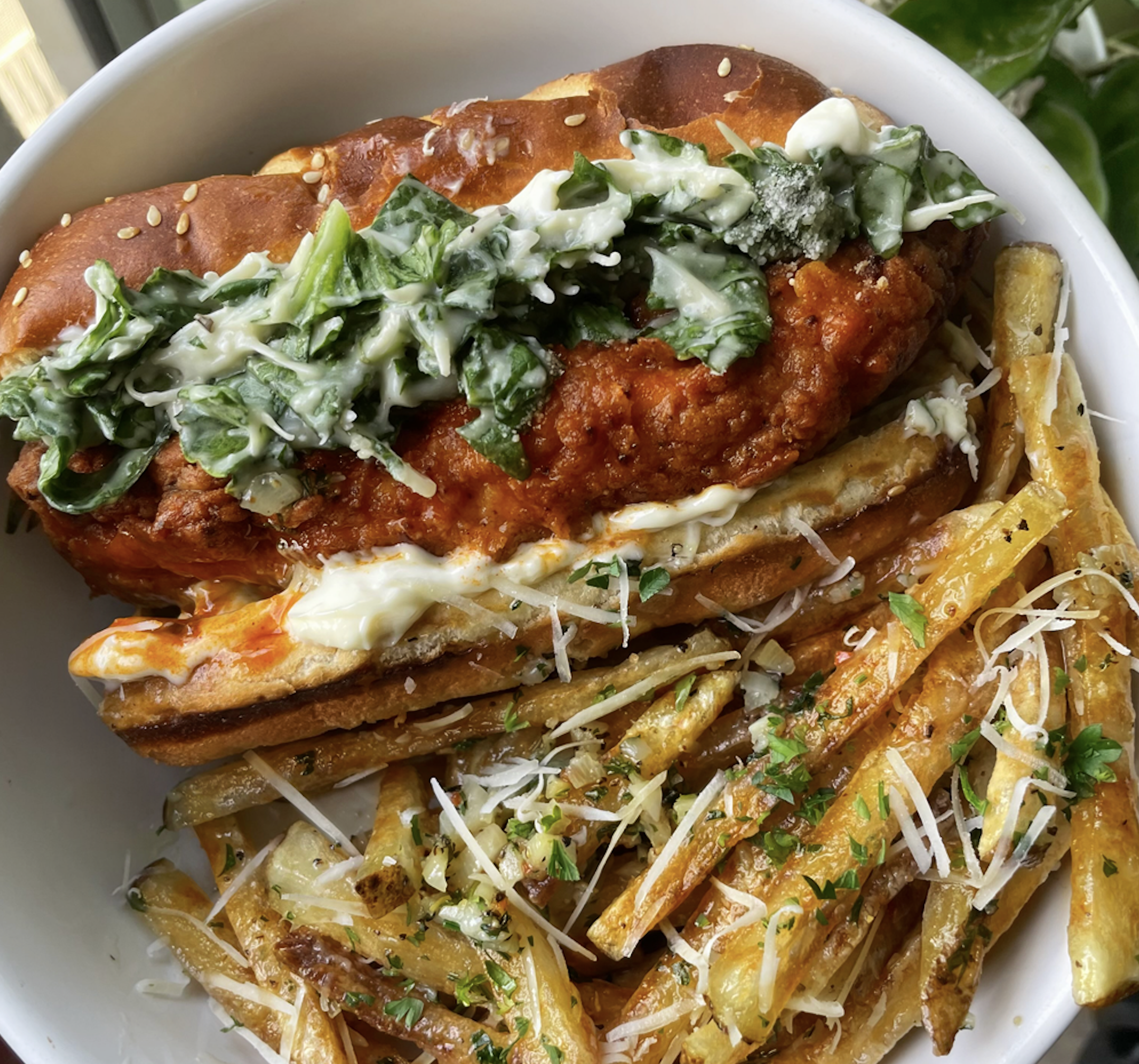 A fried chicken sandwich topped with creamy greens in a sesame bun, served with a side of seasoned fries sprinkled with grated cheese and herbs, all in a white bowl.