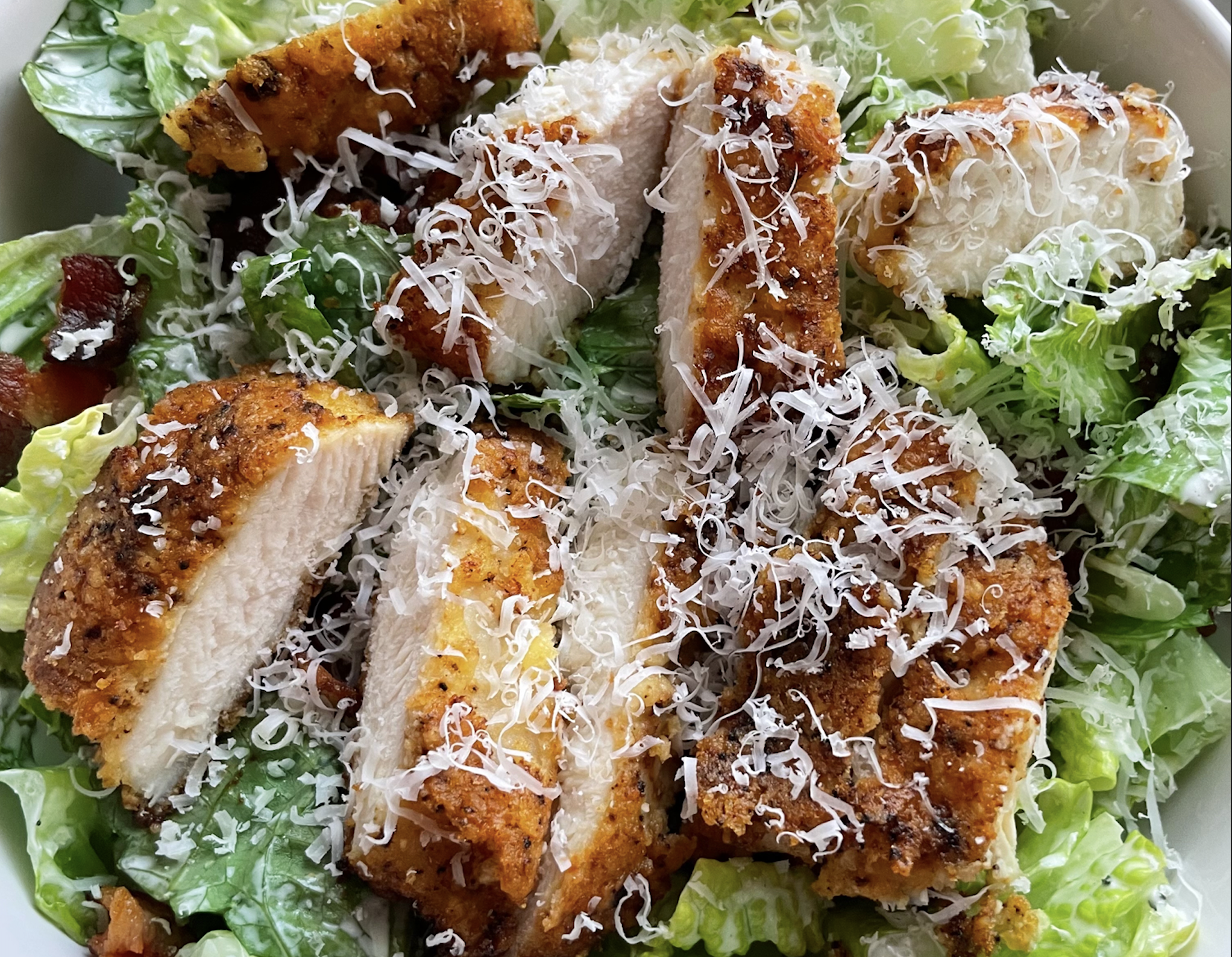 Sliced crispy breaded chicken breast on a bed of romaine lettuce, topped with shredded cheese and bacon bits.