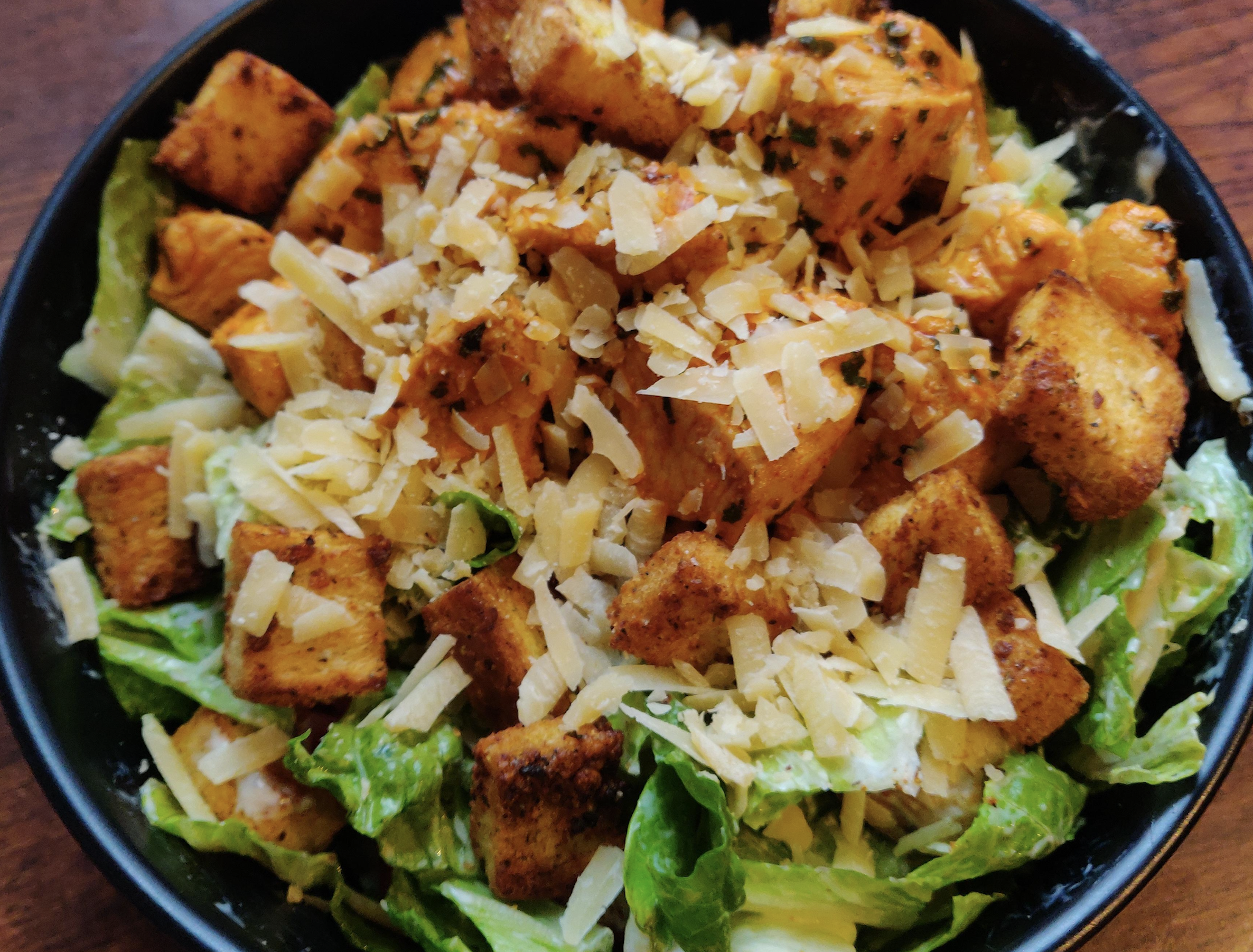 A close-up of a salad with romaine lettuce, seasoned croutons, shredded cheese, and a sprinkling of herbs in a black bowl on a wooden surface.